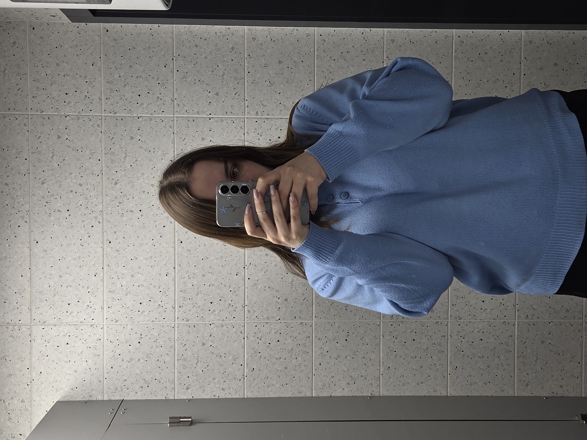 A person is holding a smartphone in front of their face, likely taking a selfie, in a bathroom. The person has shoulder-length brown hair, and is wearing a light blue sweater with buttons. The background consists of a tiled wall with a speckled pattern. Part of a gray metal structure is visible at the bottom of the image, likely a bathroom stall. The location and city are unspecified.