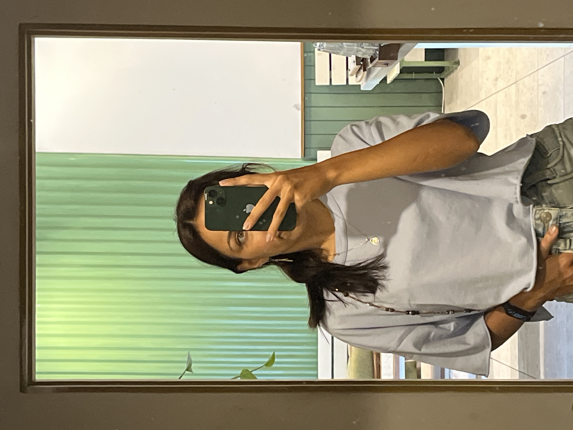 A person is taking a selfie in a mirror. The mirror shows a white board, green wall, and some decor. The person is wearing a light blue shirt, light gray pants, and a necklace, along with a wrist band. The location appears to be Kuta Selatan, Indonesia, based on common knowledge of typical indoor environments.