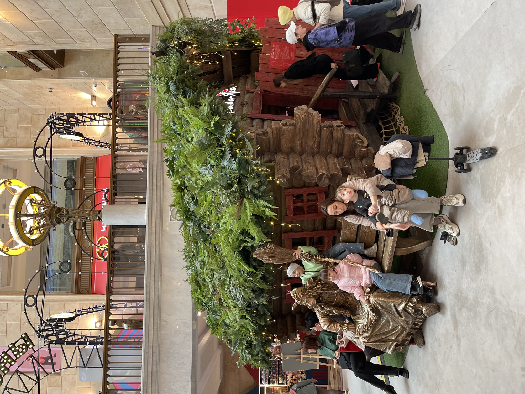 A rotated wide-angle shot shows a group of people in the Central Children's Store (also known as the Central Department Store), located in Moscow, Russia, posing on a wooden set designed to look like a rustic house or building.
The set is adorned with greenery and decorative lights. Several people are on the set, including children and adults. Some are sitting on what appears to be a wooden bench or platform. To the left of the scene is an elaborate overhead display featuring decorative elements and lighting fixtures.