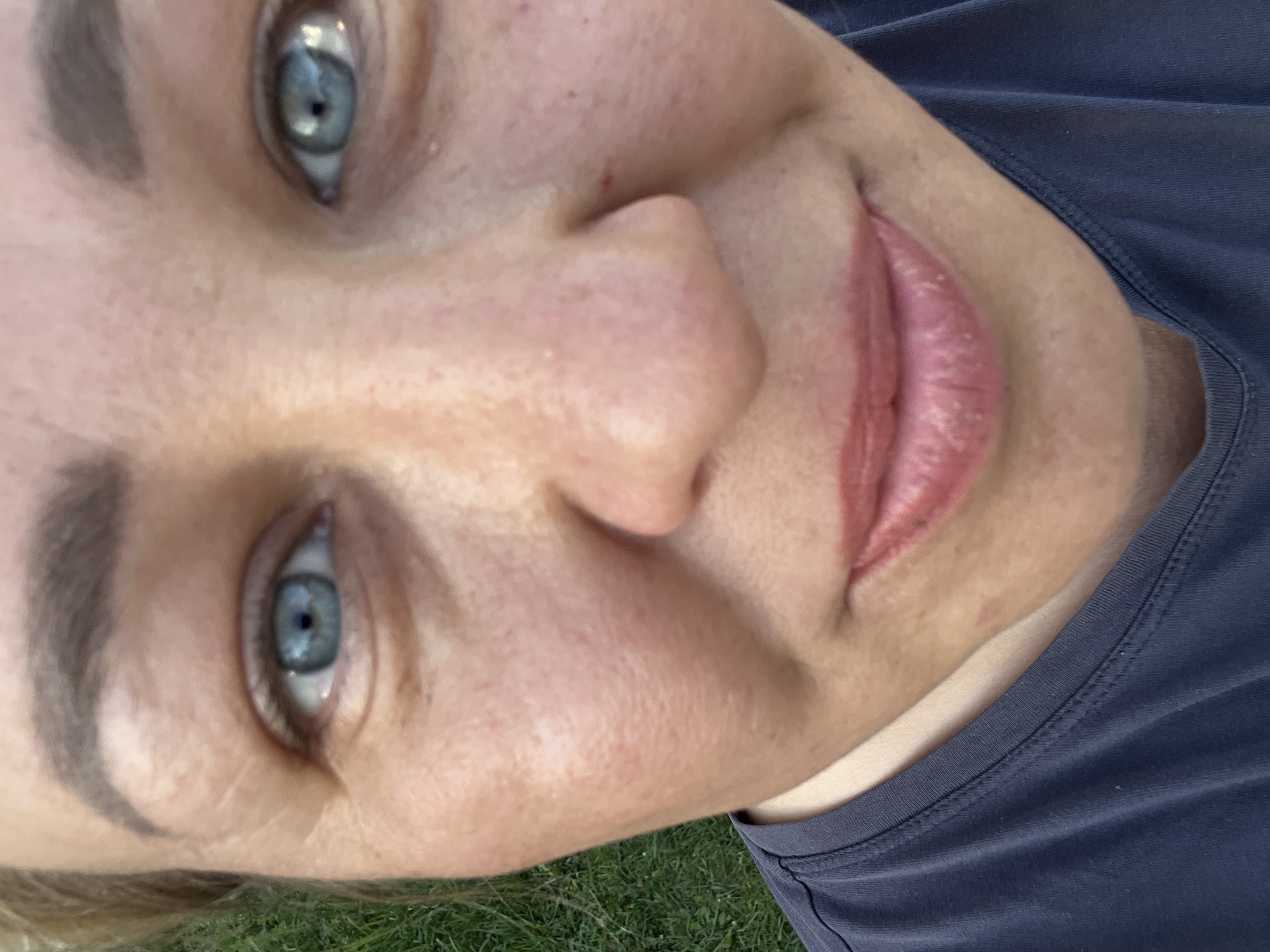 A close-up, slightly rotated image focuses on a woman's face. Her eyes are light blue. Her skin tone is fair with freckles. Her lips appear naturally pink and slightly parted. She is wearing a dark blue t-shirt. The background shows a small patch of green grass in the corner of the image. The location is unknown, and I cannot verify the city of Chaikovsky, Russia.