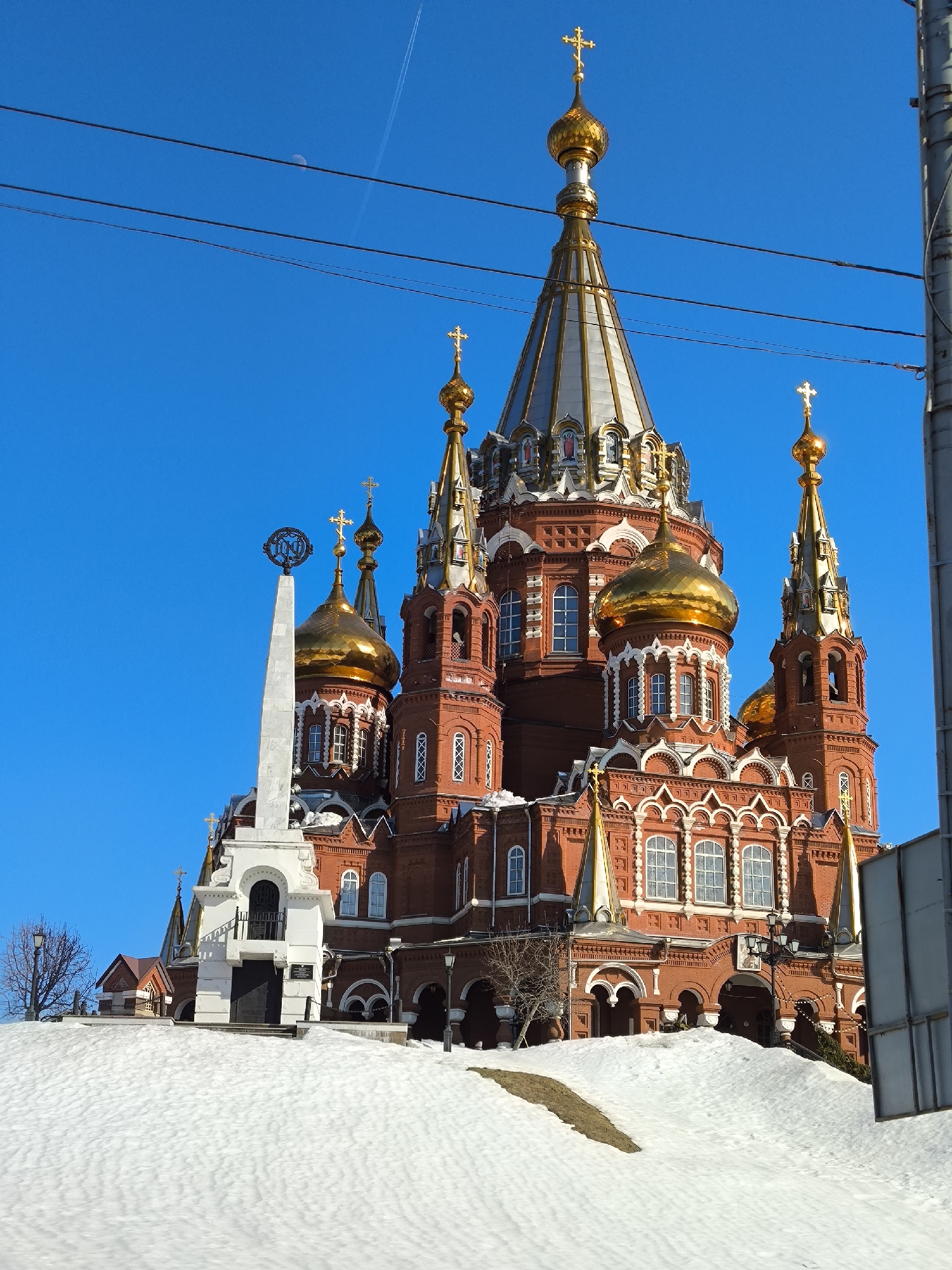 The image displays the Alexander Nevsky Cathedral in Izhevsk, Russia, under a clear blue sky. The cathedral is constructed from red brick, featuring white decorative elements around its numerous arched windows and architectural details. It is characterized by multiple golden onion domes, each topped with a golden Orthodox cross, and a large central conical dome with a silvery-gold finish, also surmounted by a golden cross.
The foreground consists of a snow-covered incline, showing undulating textures and patches of exposed brown earth. To the left of the cathedral, a tall, white obelisk-like monument with a dark, intricate circular emblem at its peak stands. A bare-branched deciduous tree is partially visible further left, behind the snow. Streetlights are visible near the base of the cathedral. On the far right, a gray metallic pole or vertical structure is partially cropped. Overhead, two dark horizontal utility wires stretch across the sky. A crescent moon is discernible in the upper left quadrant of the sky, and a faint white aircraft contrail is present in the upper right. No individuals or animals are present in the scene.