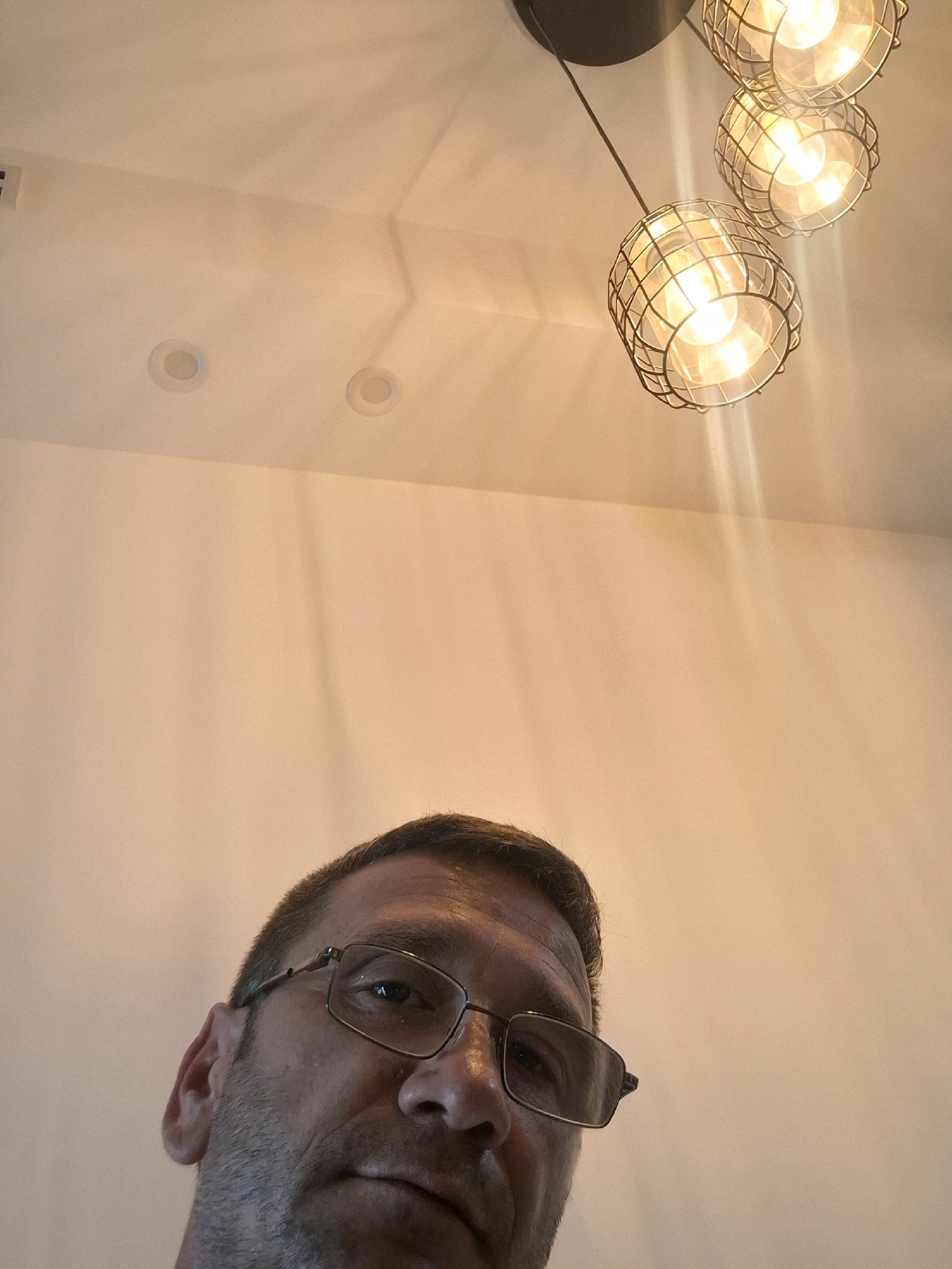 An individual, identified as male, is captured in a close-up, low-angle selfie. The man is wearing eyeglasses with dark rims and appears to be looking upwards. The background consists of a plain, light-colored wall and ceiling. A modern chandelier with three caged light bulbs is visible hanging from the ceiling, emitting a warm glow. Two recessed ceiling lights are also present. The image does not provide any specific information about the city of Ashkelon, Israel, or any other location.