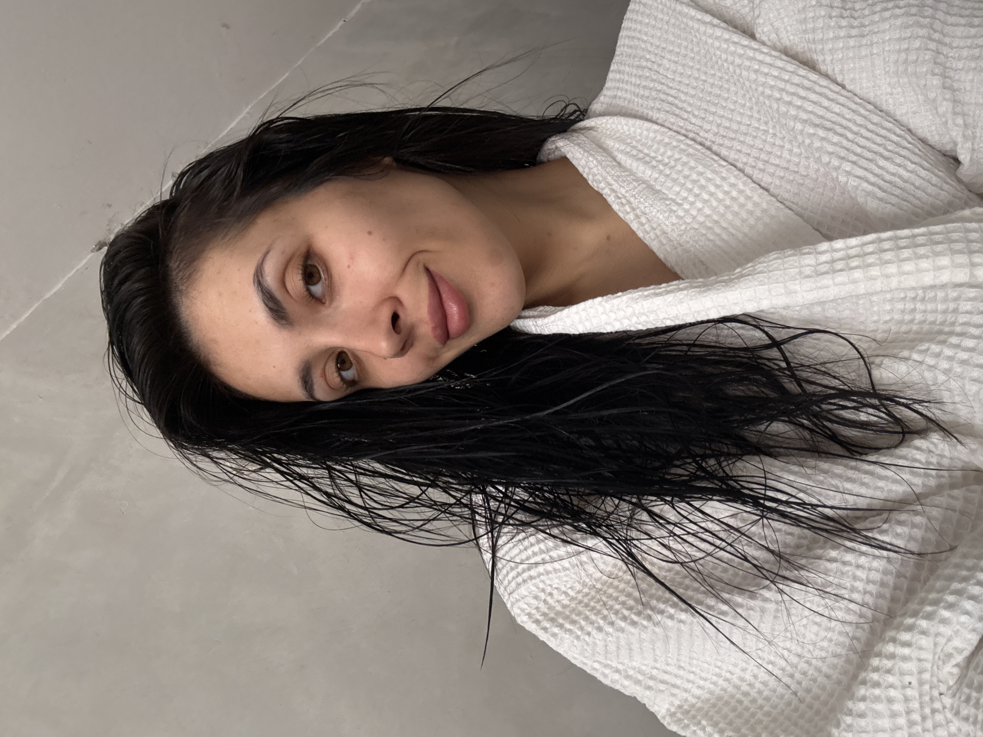The image shows a close-up, front-facing selfie of a young woman with long, dark, wet hair. She is wearing a white waffle-knit bathrobe. Her hair is styled to one side, falling over her shoulder and onto the robe. She is looking at the camera with a slight smile. The background appears to be a textured, neutral-colored wall. The lighting is soft and natural, with the woman's face well-illuminated. The image is tightly cropped, focusing on the woman's face and upper body. There are no discernible landmarks or elements that would indicate a specific location within the city of Votkinsk, Russia.