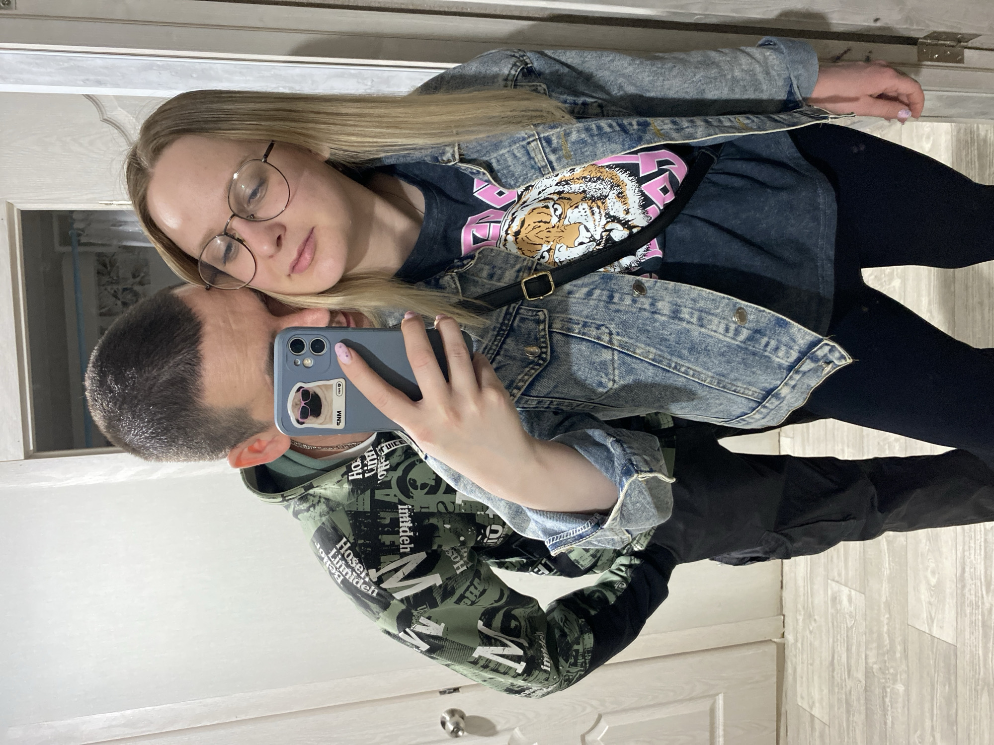 A photo taken in an interior of Unknown, Unknown, shows a woman with long blonde hair and glasses leaning her head on the shaved head of a man taking a selfie in a mirror. The woman wears a denim jacket over a shirt with a tiger design and black pants. The man wears a patterned hoodie. A phone with a sticker on the back is visible, obscuring the man's face. The background includes a door and a wall.