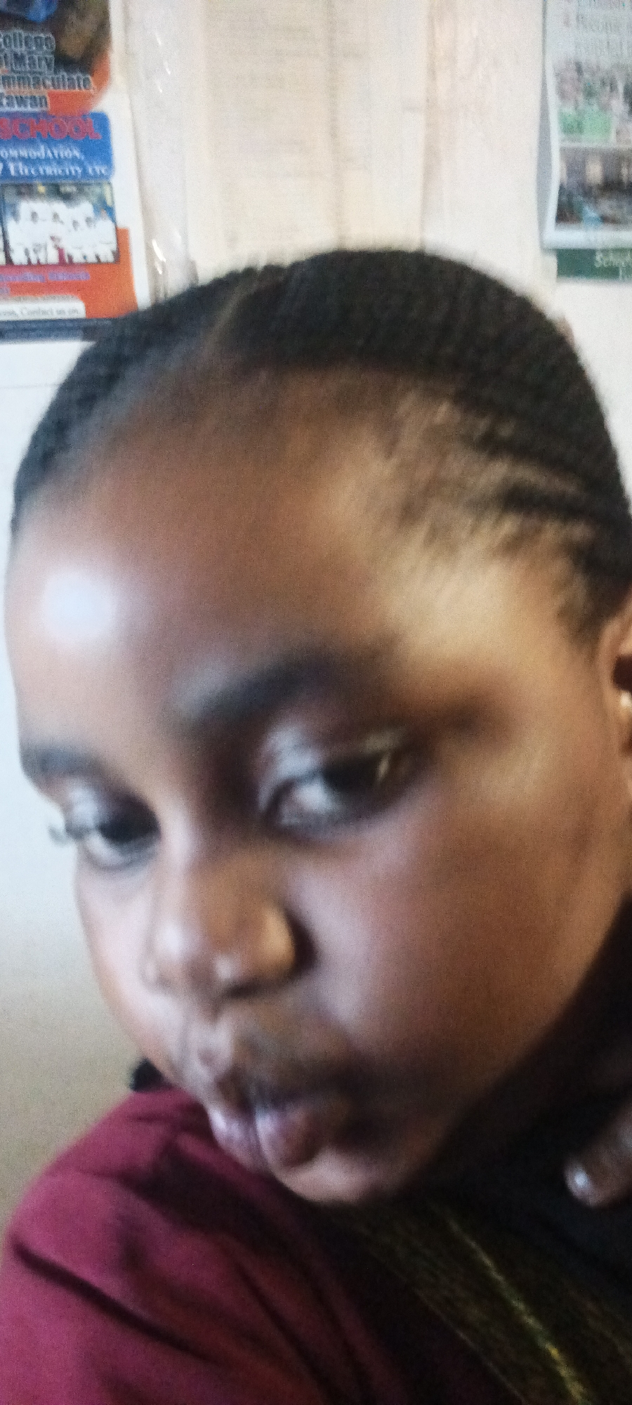 A close-up shot of a person's face, with the person looking down and slightly to the left. The person has dark skin and a hairstyle featuring braids that are laid flat against the head. The person's lips are pursed slightly. A portion of a dark red shirt is visible on the lower left side of the frame. In the blurred background, there are papers or posters hanging on a wall, some of which contain text and images related to a school. The location is in an unidentified city.