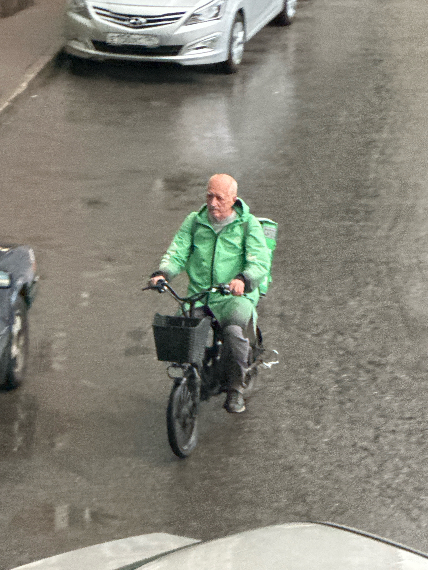 A man with a bald head and a light mustache rides a bicycle on a wet, asphalt road. He is wearing a bright green hooded jacket over a grey shirt and dark pants. A black basket is attached to the front of his bicycle, and a green delivery bag is strapped to the back of the bicycle. The man is looking straight ahead and appears to be in motion. In the background, a silver car is parked on the left side of the road. The road surface is reflective, indicating recent rain or moisture. The scene appears to be in a city, likely in Russia given the context, possibly on a street or in a parking area.