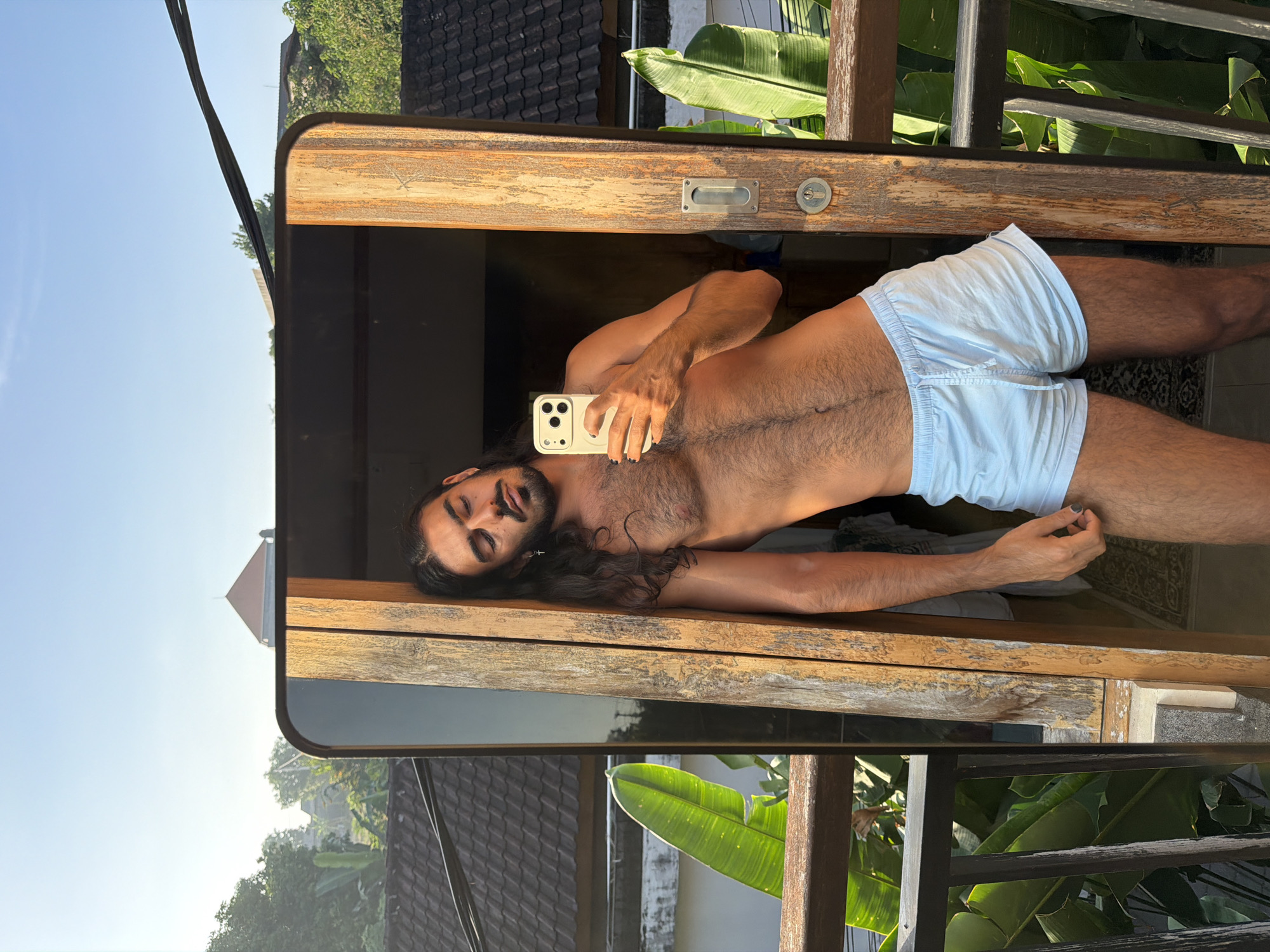 In North Kuta, Indonesia, a shirtless man with long, dark hair and a beard leans against a wooden doorframe, holding a smartphone to take a mirror selfie. He is wearing light blue shorts. The background shows a balcony with a railing, lush green foliage, and rooftops. The scene is illuminated by the warm light of a sunrise or sunset.