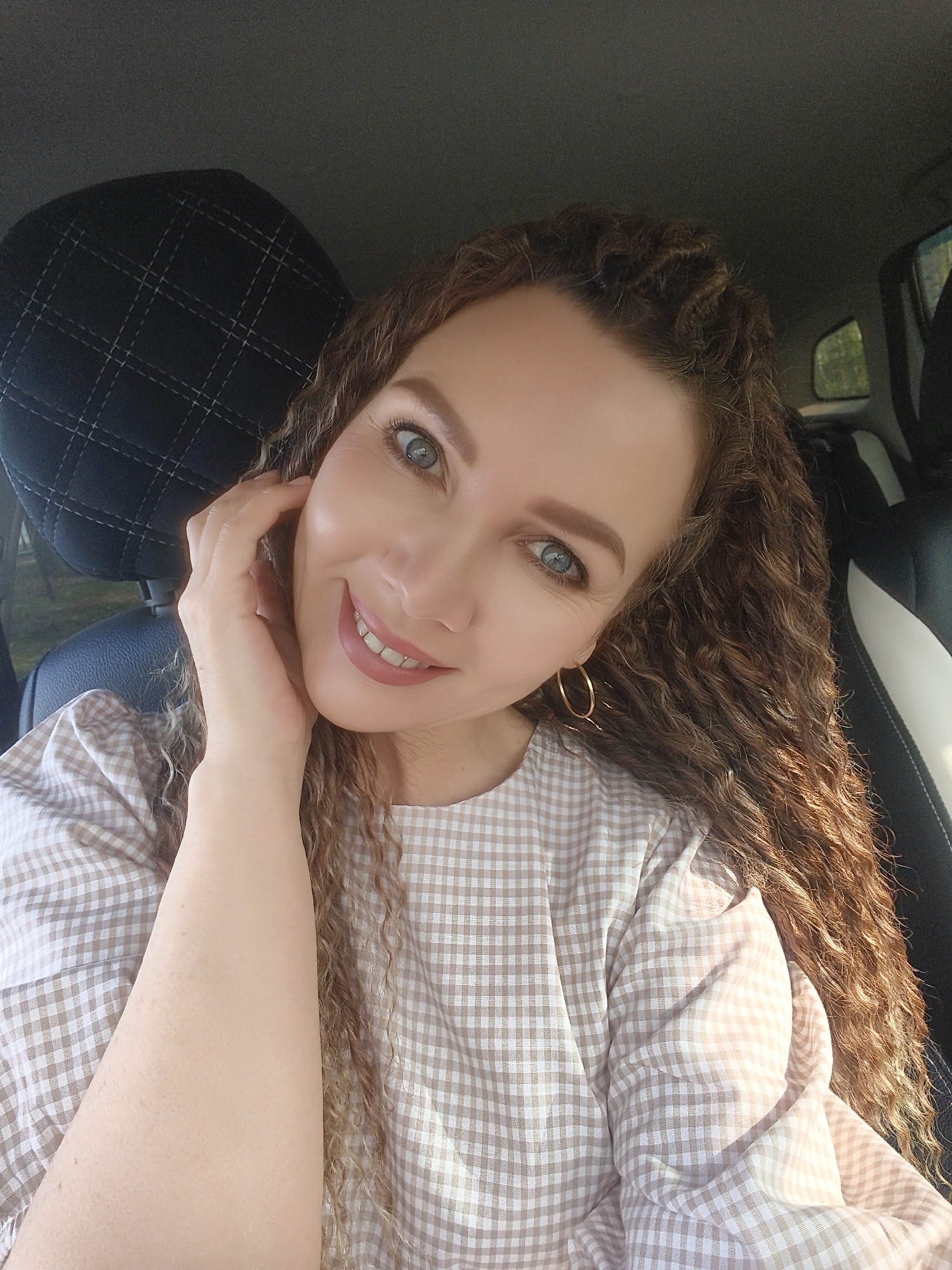 A medium shot shows a woman smiling and holding her hand to her face in what appears to be the front seat of a car. The woman has curly brown hair, light skin, and blue eyes. She is wearing gold hoop earrings and a light brown and white checkered shirt. A black headrest is visible behind her left shoulder, and the interior of the car appears to be black with some white trim. The exact location in the city of Unknown, Unknown cannot be determined from this image.