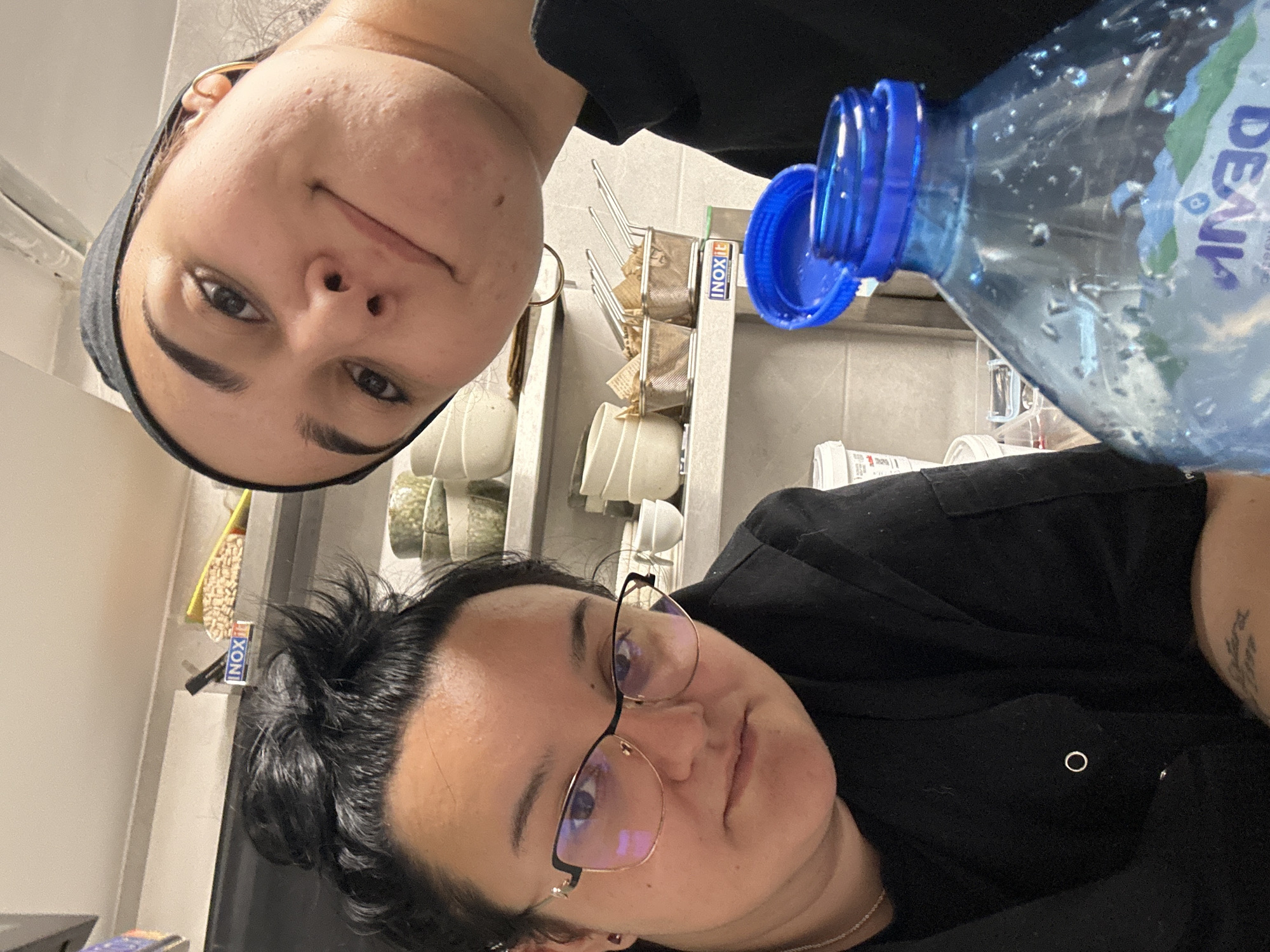 Two women are in an indoor environment. The woman on the left has fair skin, dark hair tucked under a headband, and is wearing earrings. The woman on the right has dark hair, wears glasses, and is also wearing earrings and a necklace. Both are wearing dark clothing. A partially empty bottle of "DEVIN" water is visible in the foreground. In the background, there is a shelf with what appears to be food storage containers and organizational supplies labeled "INOXIT." The scene is likely in a kitchen or storage area. There is no specific indication that the image was taken in Burgas, Bulgaria.