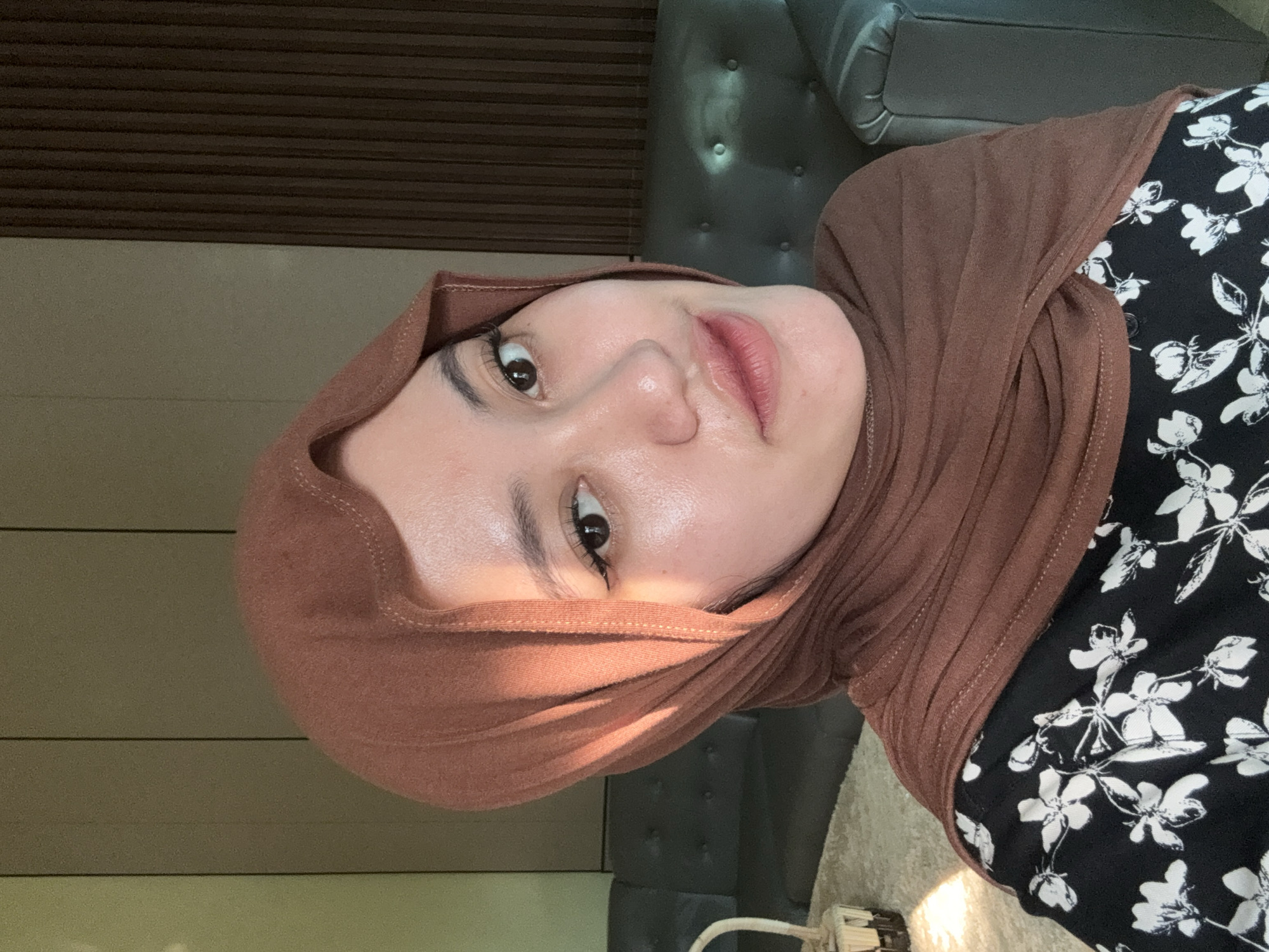 A woman wearing a brown hijab and a black shirt with white floral patterns is positioned in the foreground, occupying the central visual space of the image. Her face is well-lit, showcasing her features. In the background, a dark grey leather sofa with tufted buttons is partially visible behind her shoulder. To the left, there's a wall with horizontal wooden panels above a light beige wall. The scene appears to be indoors, possibly in a residential or commercial interior. The location is Gadingrejo, Indonesia.