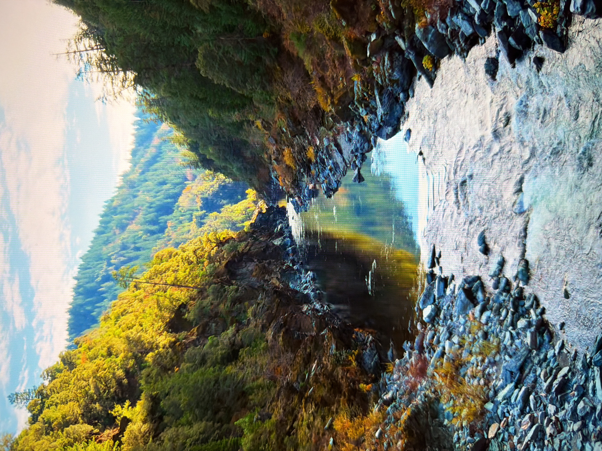 The image depicts a serene, natural landscape featuring a river flowing through a gorge flanked by steep, tree-covered hills. The foliage on the hills displays autumnal colors, with vibrant yellows and greens mixed with darker evergreen hues. The river's surface reflects the sky and the surrounding trees, creating a sense of depth and tranquility. The banks of the river are lined with rocks and sparse vegetation. The sky above is partly cloudy, with a bright, hazy quality suggesting either sunrise or sunset. The scene is devoid of any human presence or man-made structures, presenting an untouched natural environment. This image does not show any location in the city of Kyiv, Ukraine.