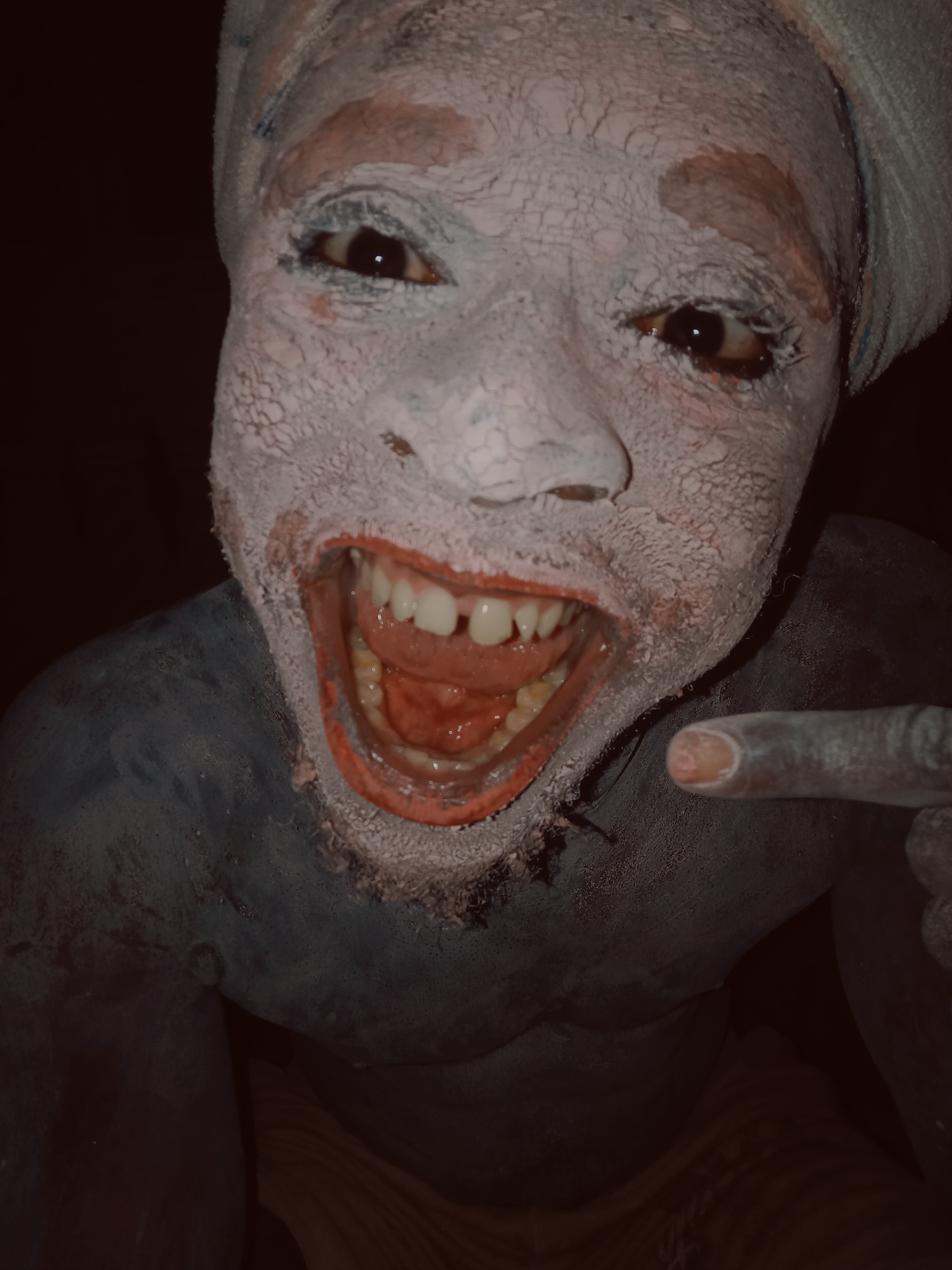 A person is covered in white and blue paint, with red around the mouth. The person is pointing a finger towards the viewer and has a wide, toothy smile. The person is wearing a white head wrap. The background is dark. The location, Onitsha, Nigeria, is assumed based on the cultural context of such body paint rituals.