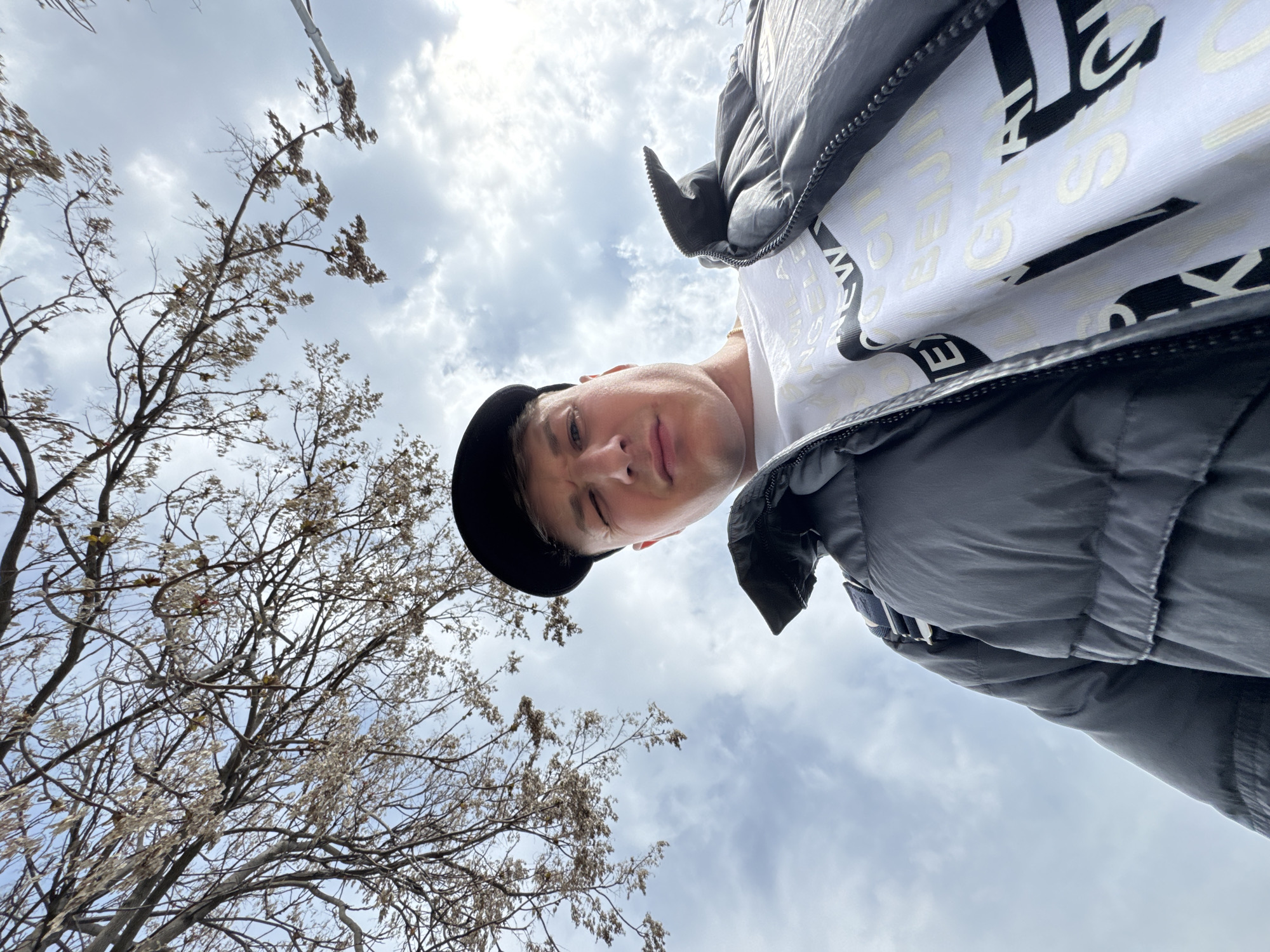 The image shows a low-angle, upward-facing selfie of a person wearing a black cap, a white t-shirt with black lettering, and a grey puffer jacket. The person's face is partially visible, with a slightly squinting gaze. The background features a tree with bare branches on the left side and a cloudy sky filling the majority of the frame. There is no definitive evidence to confirm the location is in Plovdiv, Bulgaria.
