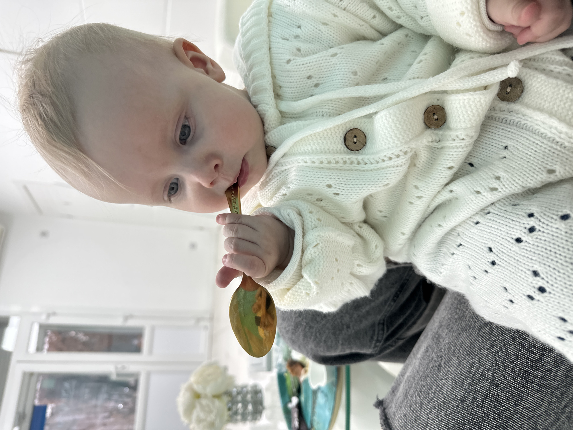 An infant with light blonde hair and blue eyes is depicted in a brightly lit, modern interior space. The infant, wearing a cream-colored knitted cardigan with a hood, wooden buttons, and an eyelet pattern, is seated on the lap of an individual whose dark gray denim-clad legs are visible. The infant's right hand holds a golden-colored spoon, the tip of which is in the infant's mouth. Text partially visible on the spoon's handle reads "England 187". In the blurred background to the left, a turquoise round tray is present on a glass-topped table, holding a small white dish with food items and a vase containing white flowers. The far background features white walls and two white doors with vertical glass panels. The specific location in Moscow, Russia, cannot be determined from the visual information presented in the image.