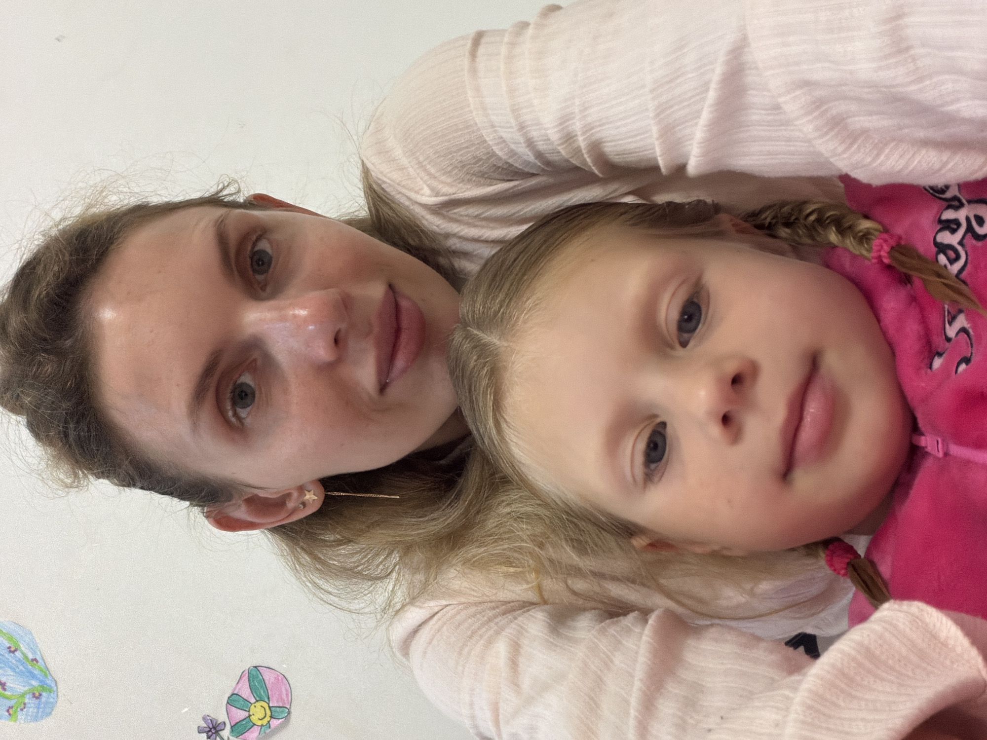 A woman and a young girl are closely posed together. The woman is on the left side, with light brown hair partially pulled back. She has fair skin, and her face is tilted slightly towards the right. The girl, positioned next to her, has blonde hair braided into pigtails, blue eyes, and fair skin. She wears a bright pink top. Both individuals are looking at the camera. The background is a light, neutral color with some simple children's drawings. The image appears to be taken indoors, in a casual setting. It is not possible to determine if the image was taken in Budva, Montenegro.