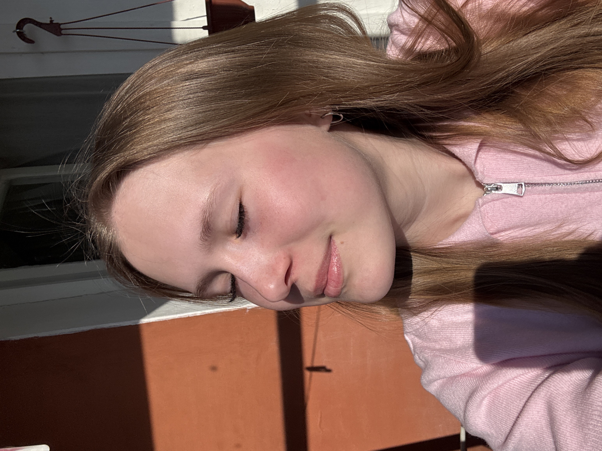 A medium, close-up, portrait shot of a fair-skinned, young person with closed eyes and long, light brown hair. They are wearing a light pink, zip-up hooded sweatshirt. The person's face is partially in shadow, with the sun casting a light across their cheek and lips. The background features a section of a white window frame, an orange-painted wall or surface, and a dark, elongated shadow. A plant holder or similar object and a wire hanger are visible in the upper left corner of the frame, silhouetted against a lighter background. The location is outdoors, with strong sunlight suggesting the image was taken during the day. There is no explicit information to conclude this was taken in Chaikovsky, Russia.