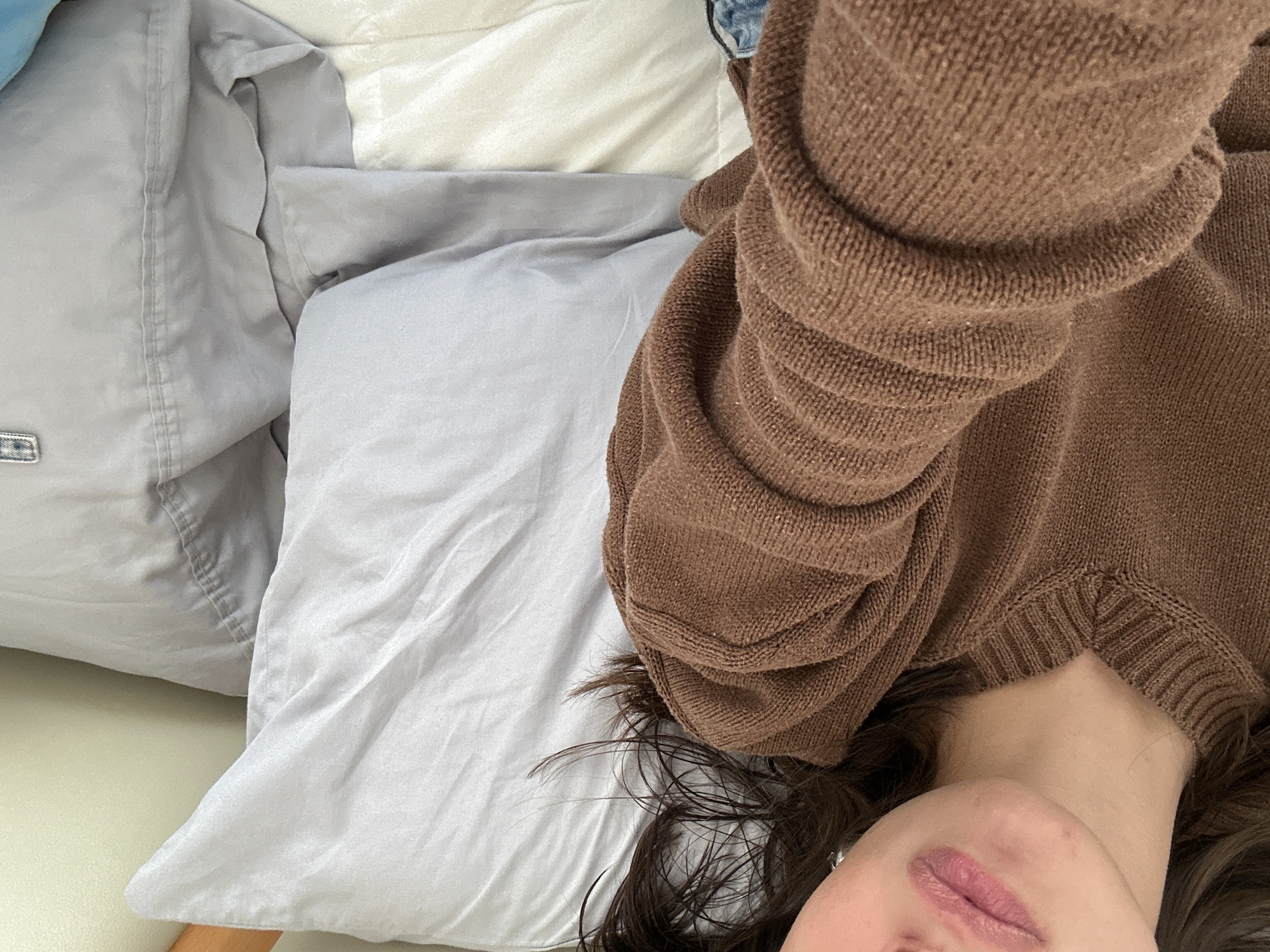 The image shows a person lying on a bed. They are wearing a brown sweater and have dark hair. The bed has gray and white bedding. The location and specific city are not discernible from the image.