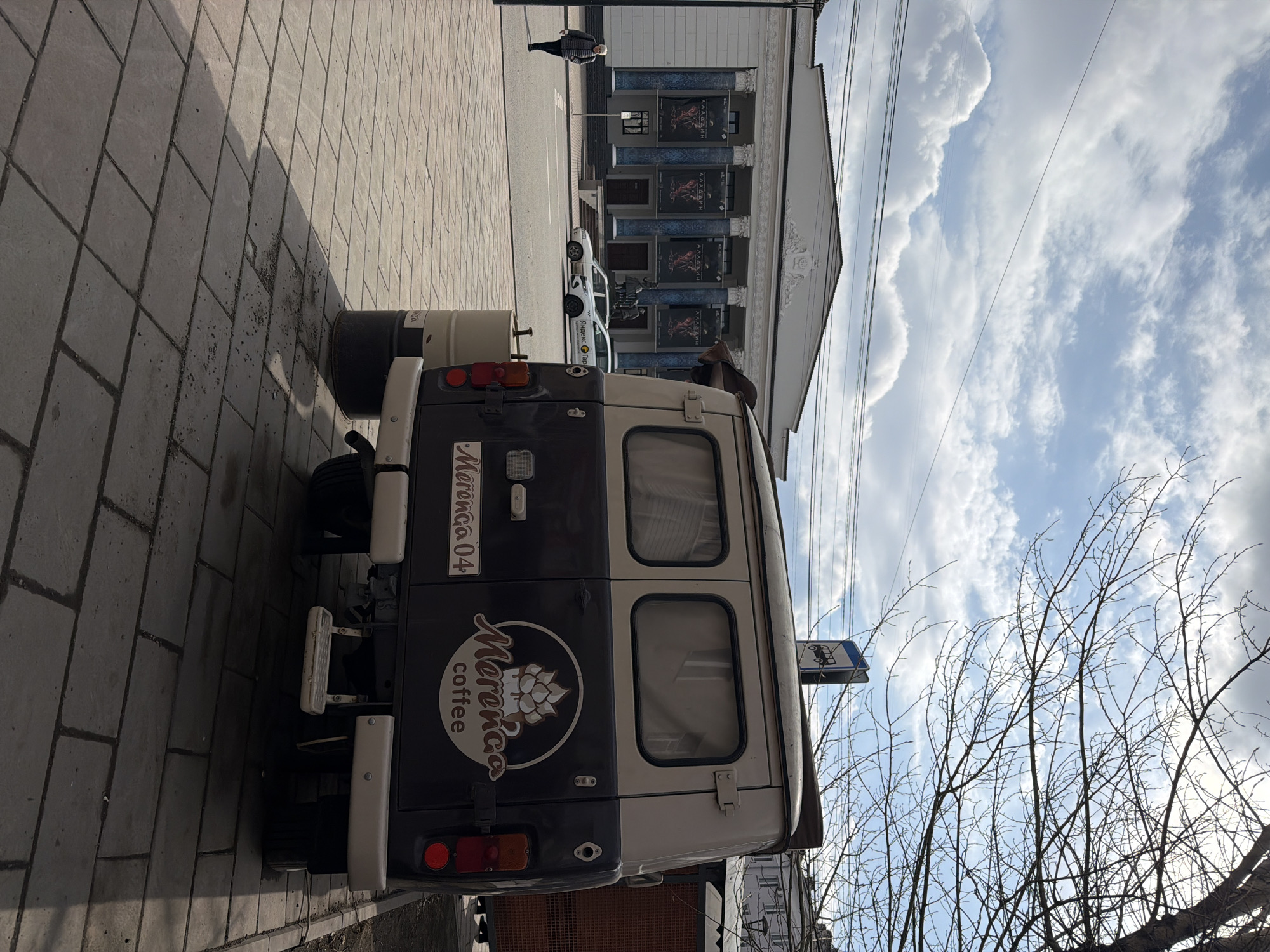The image depicts a street scene in Krasnoyarsk, Russia. A dark gray and white van, labeled "Morenco Coffee" and "Morenco 04," is parked on a paved sidewalk. A small bus stop sign is visible adjacent to the van. In the background, a building with multiple windows and decorative features is visible. A person is walking along the sidewalk in the distance. The sky is partially cloudy with bare tree branches in the foreground.