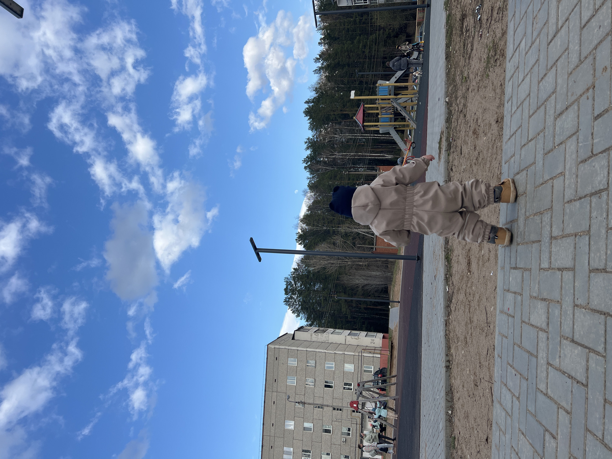A toddler in a tan jumpsuit and blue hat stands on a grey brick sidewalk. Behind them is a playground, a multi-story building, and a forest. The sky is blue with white clouds. This scene is located in Noviy, Russia.