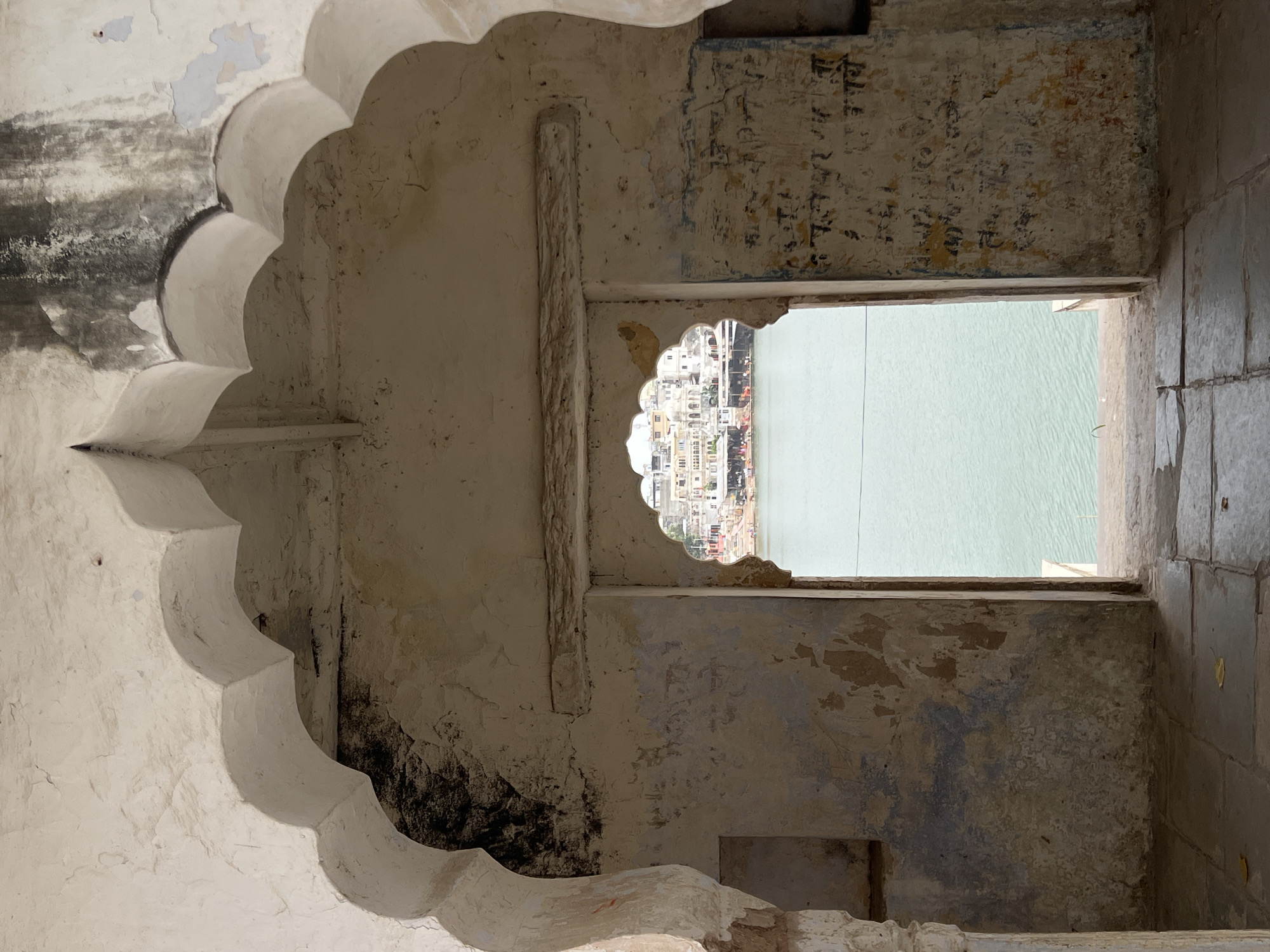 The image depicts a view through an arched doorway onto a lake with buildings in the background, likely in Pushkar, India. The foreground is dominated by the weathered stone of the structure, with arched openings and crumbling plaster walls. The walls bear traces of faded paint and what appear to be inscriptions.
Through the smaller, elaborately shaped archway of a secondary opening, a calm, turquoise body of water is visible. On the opposite side of the lake, a cluster of white buildings with numerous domes and minarets forms a skyline. The architecture suggests a temple complex or a town center common in Indian cities. The water is placid with subtle ripples, and a few boats or small vessels are visible docked along the far bank. The overall scene conveys a sense of historical architecture and a tranquil, urban lakeside environment.