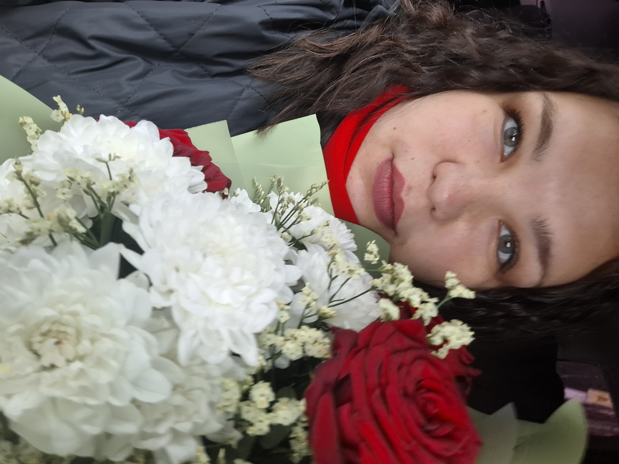 The image displays a close-up portrait of a woman holding a bouquet of flowers, taken in Chaikovsky, Russia. The woman, positioned so her head is tilted to the right, has dark, curly hair, light green or hazel eyes, and is wearing dark red lipstick. She has a neutral to slightly pleasant expression and is looking directly at the camera. Her attire includes a red garment, possibly a top or scarf, visible at her neck, and a black quilted jacket.
The bouquet, occupying the left and lower parts of the frame, features a prominent cluster of large white chrysanthemums, complemented by smaller white filler flowers. Two vibrant red roses are also visible within the arrangement, providing a contrasting color accent. The flowers are partially wrapped in light green paper. The image appears to be a selfie or a very close-up shot, with the woman and the bouquet being the central subjects. No text is visible in the media file.
