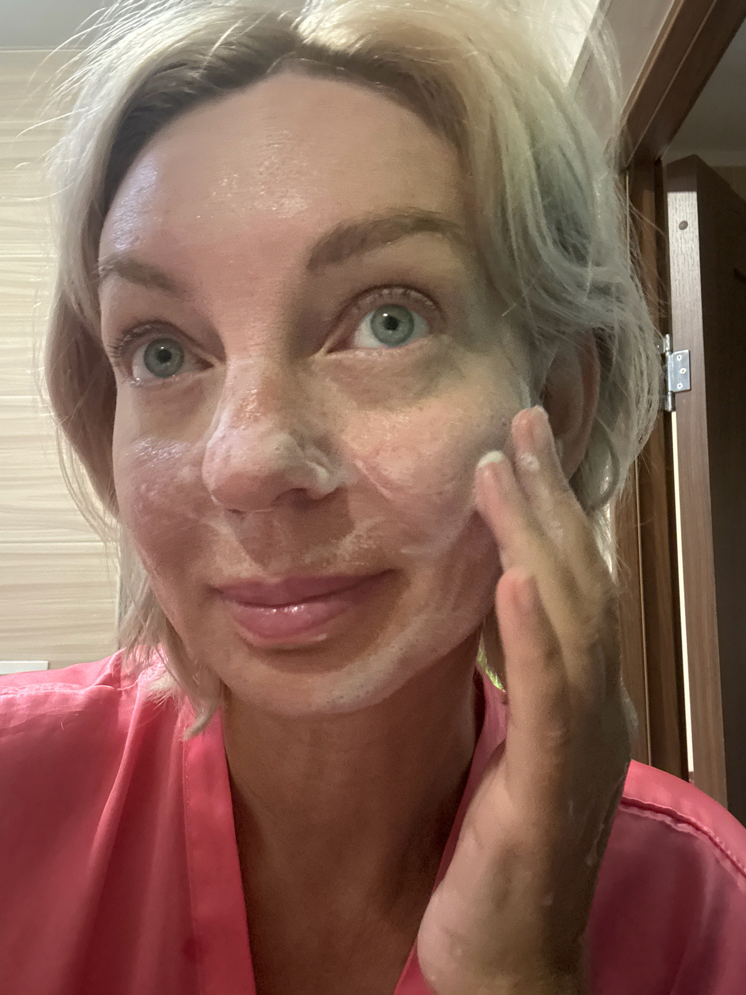 A close-up, head-and-shoulders selfie shows a woman with blonde hair and blue eyes applying a white cleansing foam to her face. She is wearing a bright pink robe. Her skin appears slightly red and irritated. The background shows a light-colored tiled wall and a wooden door frame with hinges. The image was taken in Sochi, Russia.