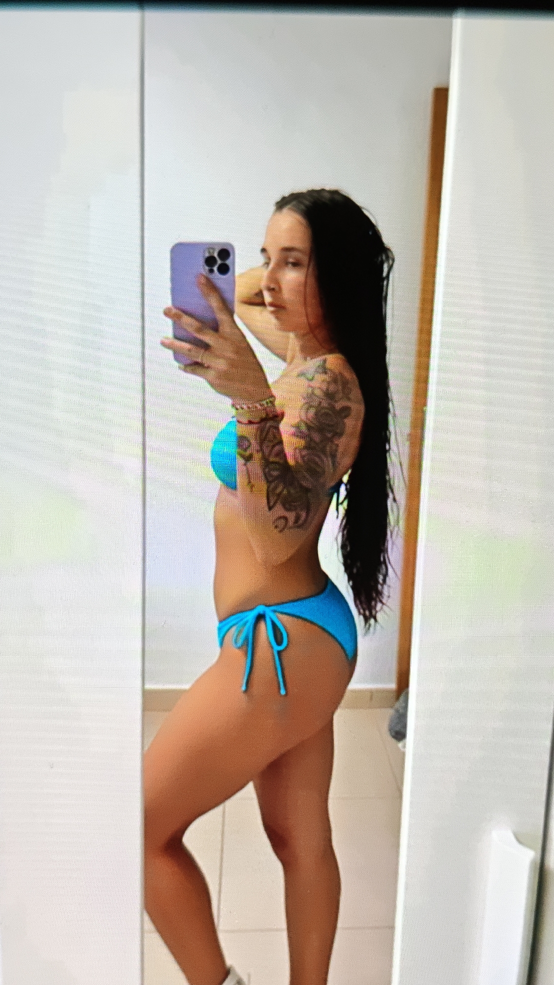 A woman is taking a mirror selfie in what appears to be a brightly lit room, possibly a bathroom, as indicated by the tiled wall visible in the reflection. She is wearing a blue bikini and has a tattoo on her right arm. She is holding a purple phone in her left hand, with her right arm behind her head. Her long, dark hair is down her back. The room's walls are white, and a full-length mirror is prominent.