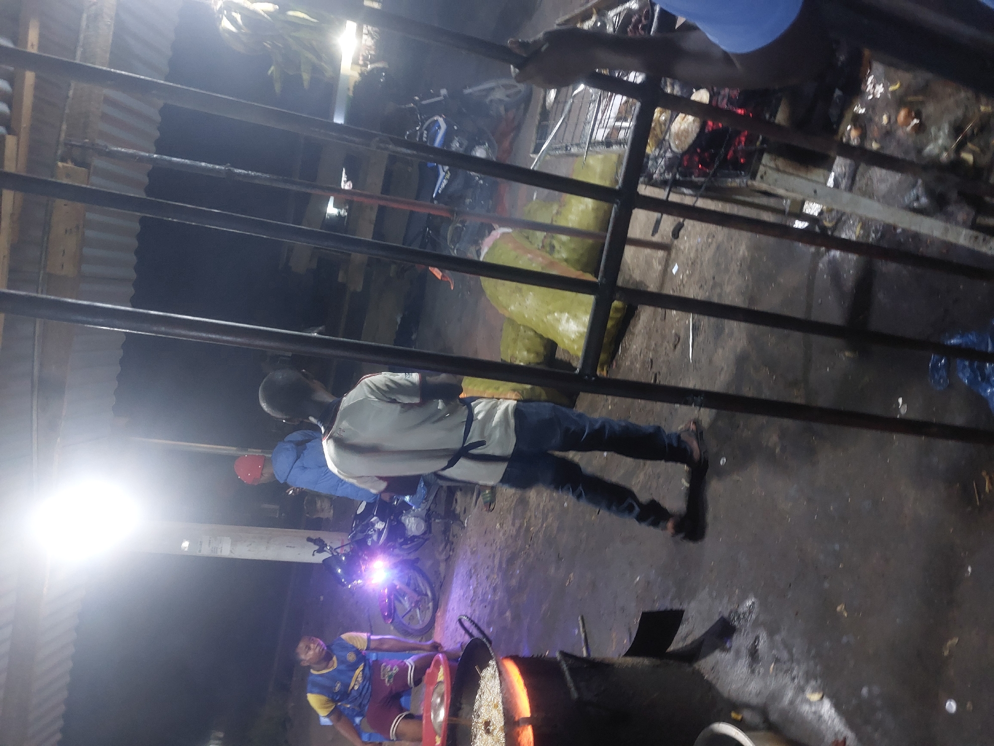 The image depicts an outdoor scene in Dar es-Salaam, Tanzania, likely at night. There is a metal grill or barrier in the foreground. Behind it, a man in a light-colored shirt and jeans is standing near a motorcycle with its headlight on. Another man in a blue and yellow shirt is sitting on the ground next to a cooking pot with glowing contents, possibly food being prepared. There is also another man with a red cap in the image. In the background, there are sacks, potentially containing food. The surroundings suggest a casual outdoor cooking and gathering area.