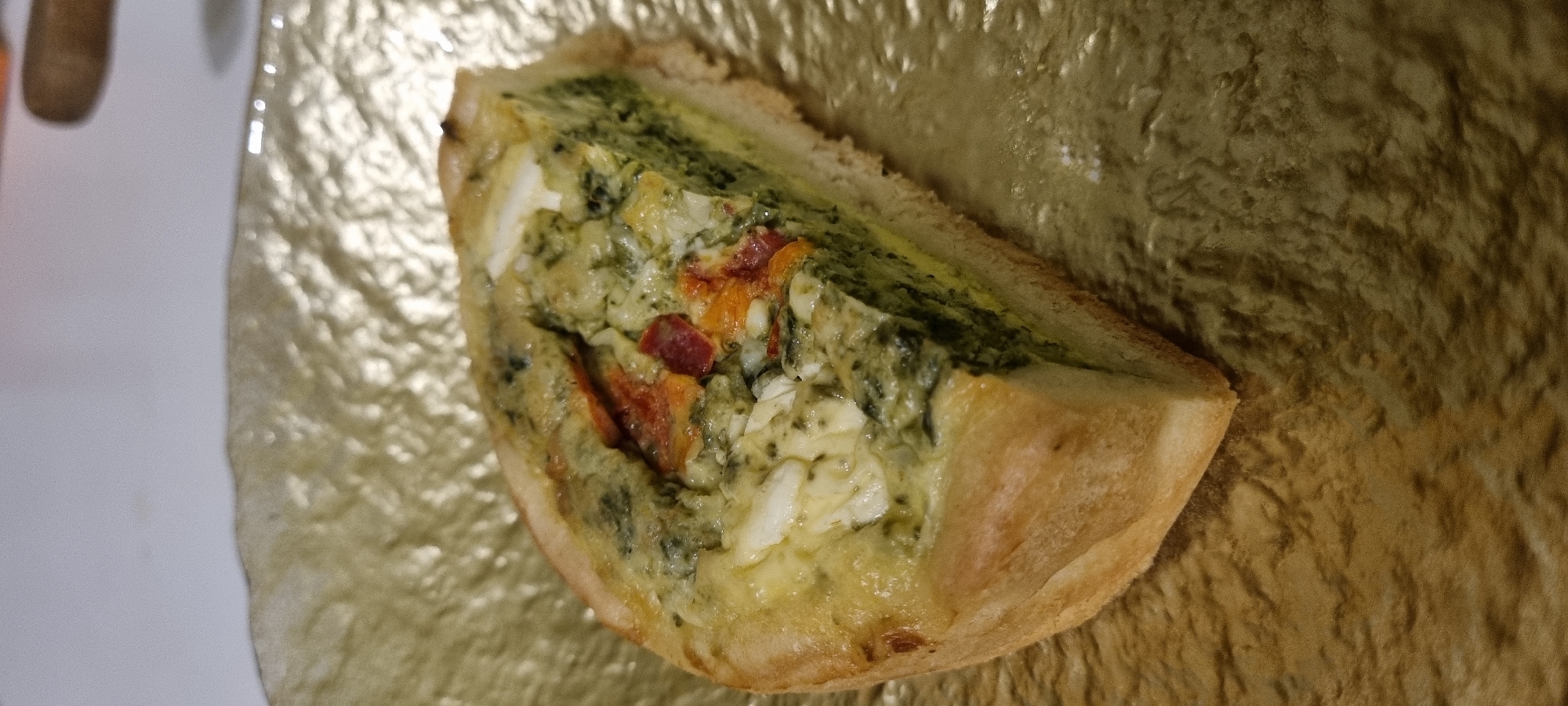 A slice of quiche with a golden crust sits on a textured golden plate. The quiche filling appears to be a mix of spinach, eggs, and possibly bacon or ham. The lighting in the image is warm and slightly diffused, highlighting the textures of both the food and the plate. The background is out of focus, suggesting a shallow depth of field. The overall impression is that of a close-up shot of a prepared dish, likely in a domestic or restaurant setting. The location in Romashkovo, Russia is not discernible from the image itself.
