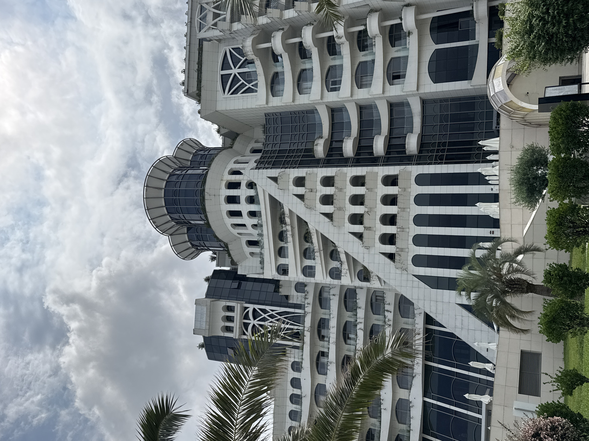 The image shows the Batumi Renaissance Hotel in Batumi, Georgia, under a cloudy sky. The hotel is a large, white, modern building with a distinctive, multi-tiered design featuring curved balconies, large dark windows, and decorative arches. Palm trees are visible in the foreground, with manicured greenery and white umbrellas on a lower level of the hotel. The architecture is ornate and complex, suggesting a luxury establishment. There are no people or visible actions in the scene.