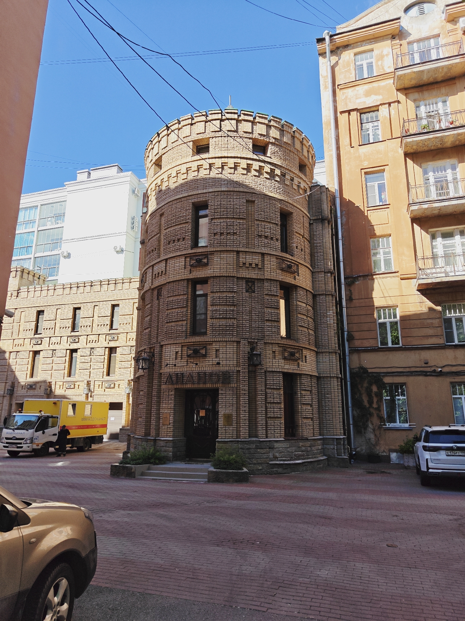 This is a daytime, eye-level, outdoor, medium-shot photograph taken in an urban setting in Saint Petersburg, Russia. The scene depicts a courtyard area between buildings.
In the foreground, the hood and front wheel of a tan-colored car are visible, parked on a red brick paved ground.
To the left of the frame, a white van with "ВОДОКАНАЛ" (Vodokanal - Water Utility) and "АВАРИЙНО-СЛУЖБА" (Emergency Service) written on its side is parked. A person in dark clothing is standing by the van, facing away from the camera.
The central and most prominent structure is a circular, multi-story building constructed of light brown bricks. This building features a decorative, crenellated parapet at the top, and a series of arched windows and decorative brickwork along its facade. A dark wooden door is visible at the base of this structure, with steps leading up to it. Above the door, text that appears to read "ATLANTIS" is visible.
To the right of the circular building is a taller, orange-colored apartment building. This building has multiple floors with balconies and windows. A downspout runs vertically along the side of this building.
In the background, a white, modern building with large windows is visible behind the circular brick structure. The sky above is clear and blue. Electrical wires are strung across the sky in the upper portion of the image. The overall impression is one of an older European city with a mix of architectural styles.