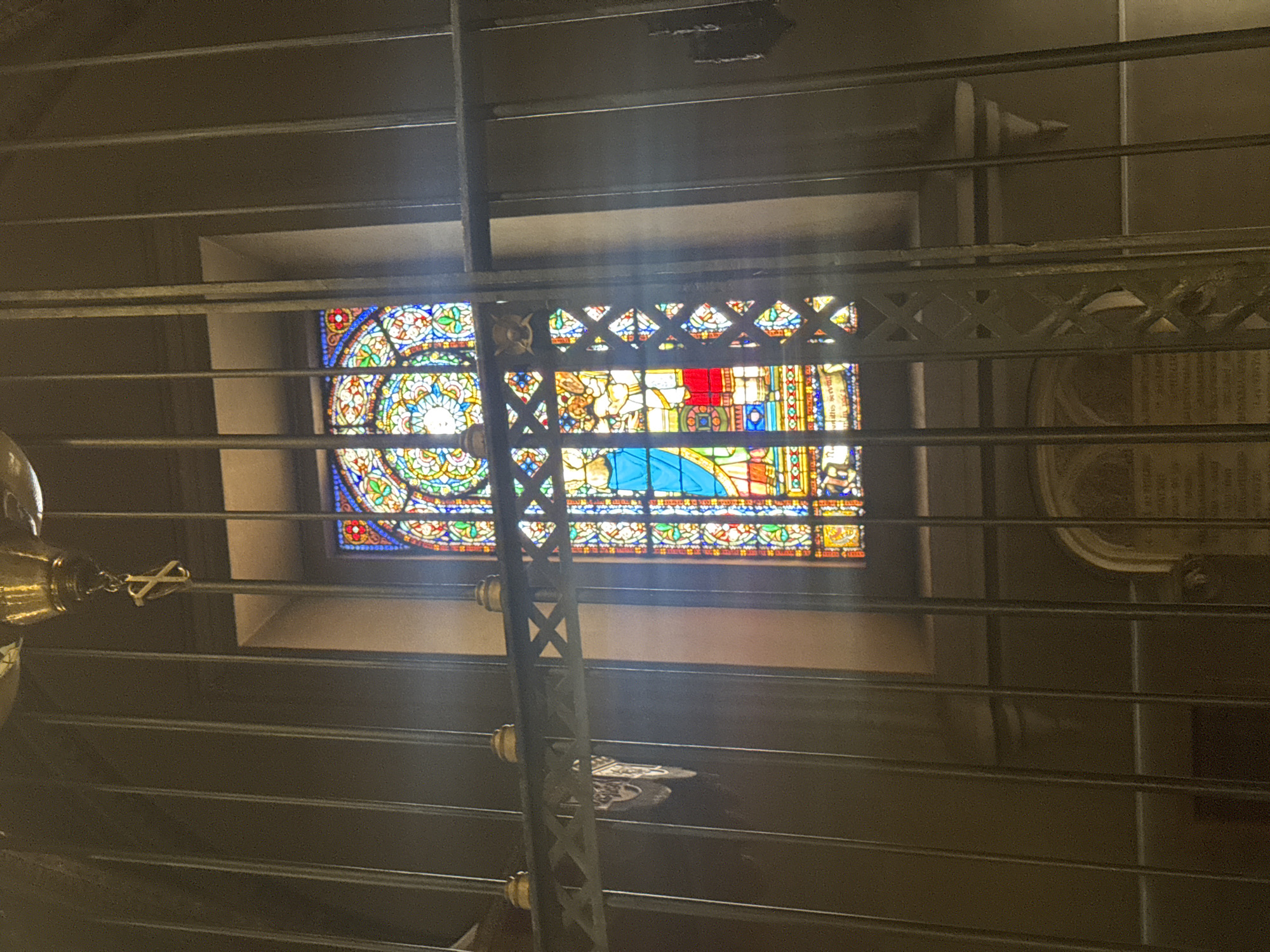The image shows a stained-glass window depicting religious figures, framed by dark metal bars. The window is arched and highly ornate, with a central figure in a blue robe. The overall setting appears to be indoors, possibly a church or a historical building, with a dimly lit environment. There are no people visible in the frame. The metal bars create a sense of enclosure or separation from the viewer. The location is identified as the Monistrol de Montserrat, Spain, though specific details within the image do not directly confirm this.