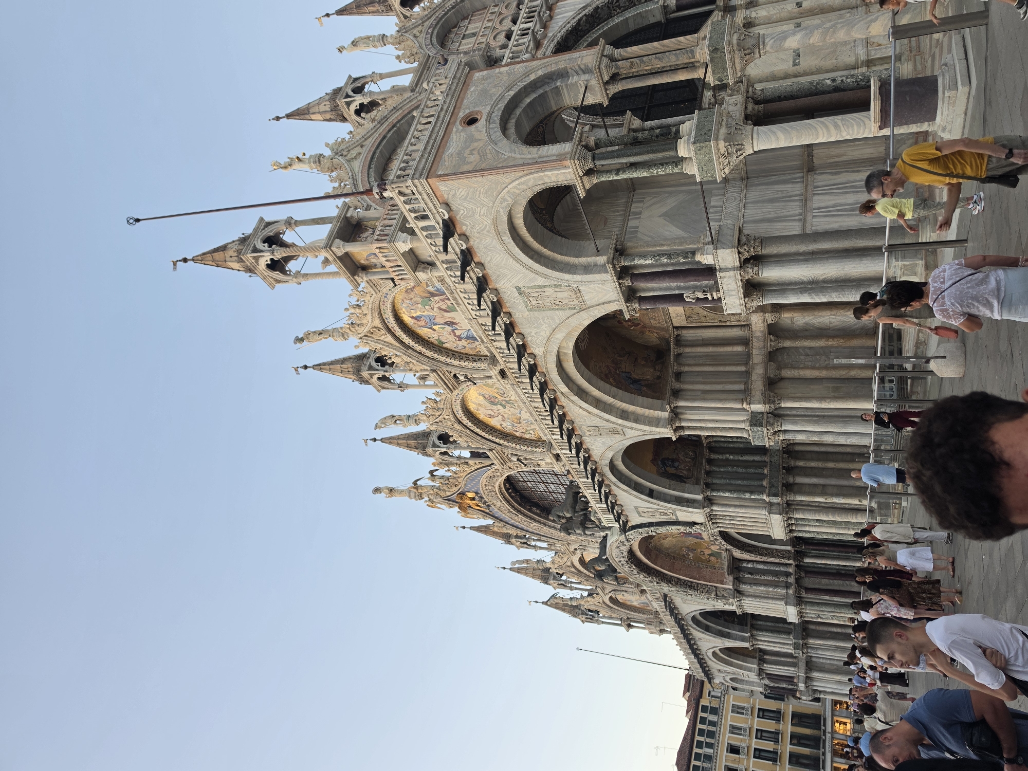 The image captures an exterior view of St. Mark's Basilica in Venice, Italy, under a clear sky. The basilica's ornate facade, characterized by multiple arches, columns, and intricate sculptures, is prominently featured. Mosaic artwork is visible above the arches. A crowd of people is milling about in the foreground and midground, some appearing to be tourists. The architectural style suggests a Byzantine influence. The lighting indicates it is likely late afternoon or early evening.