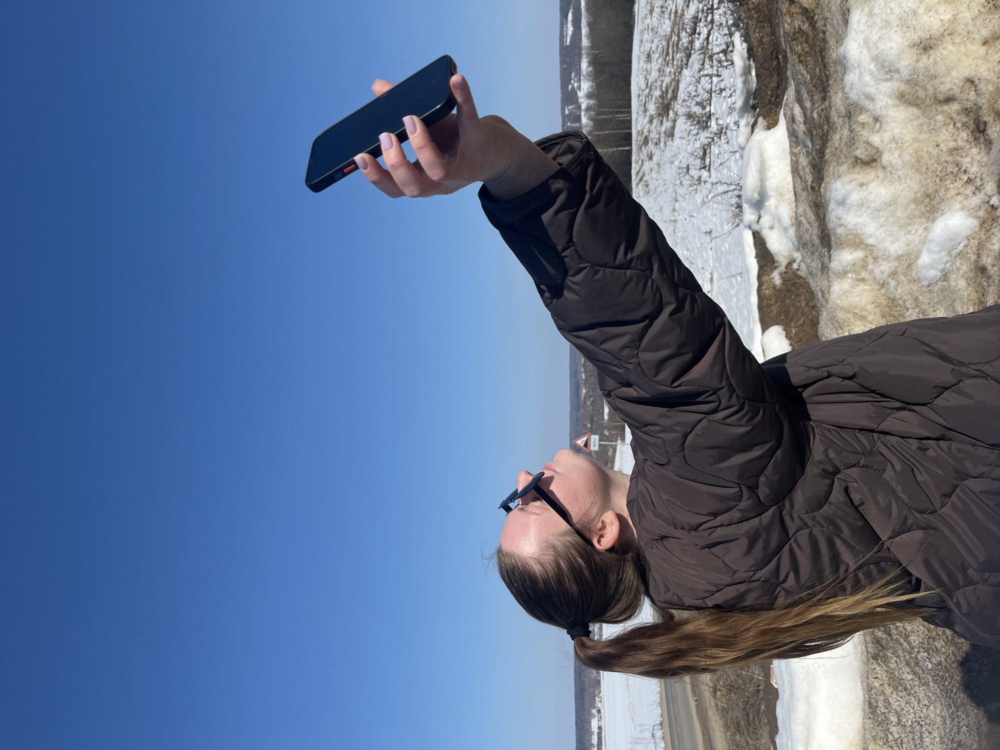 A female subject is depicted outdoors in Zubovo, Russia, under a clear blue sky. She stands in profile, gazing upwards, with her right arm extended overhead, holding a dark smartphone with a red side button. Her long brown hair is pulled into a ponytail, and she wears sunglasses and a dark brown quilted jacket. The immediate ground displays patches of melting snow and brown earth. A paved road is visible on the left, slightly below the subject's position. In the mid-ground, a partially visible red-bordered triangular road sign indicates "400m." The background consists of a winter-to-spring landscape featuring snow-covered fields, hills, and a distant line of bare trees.