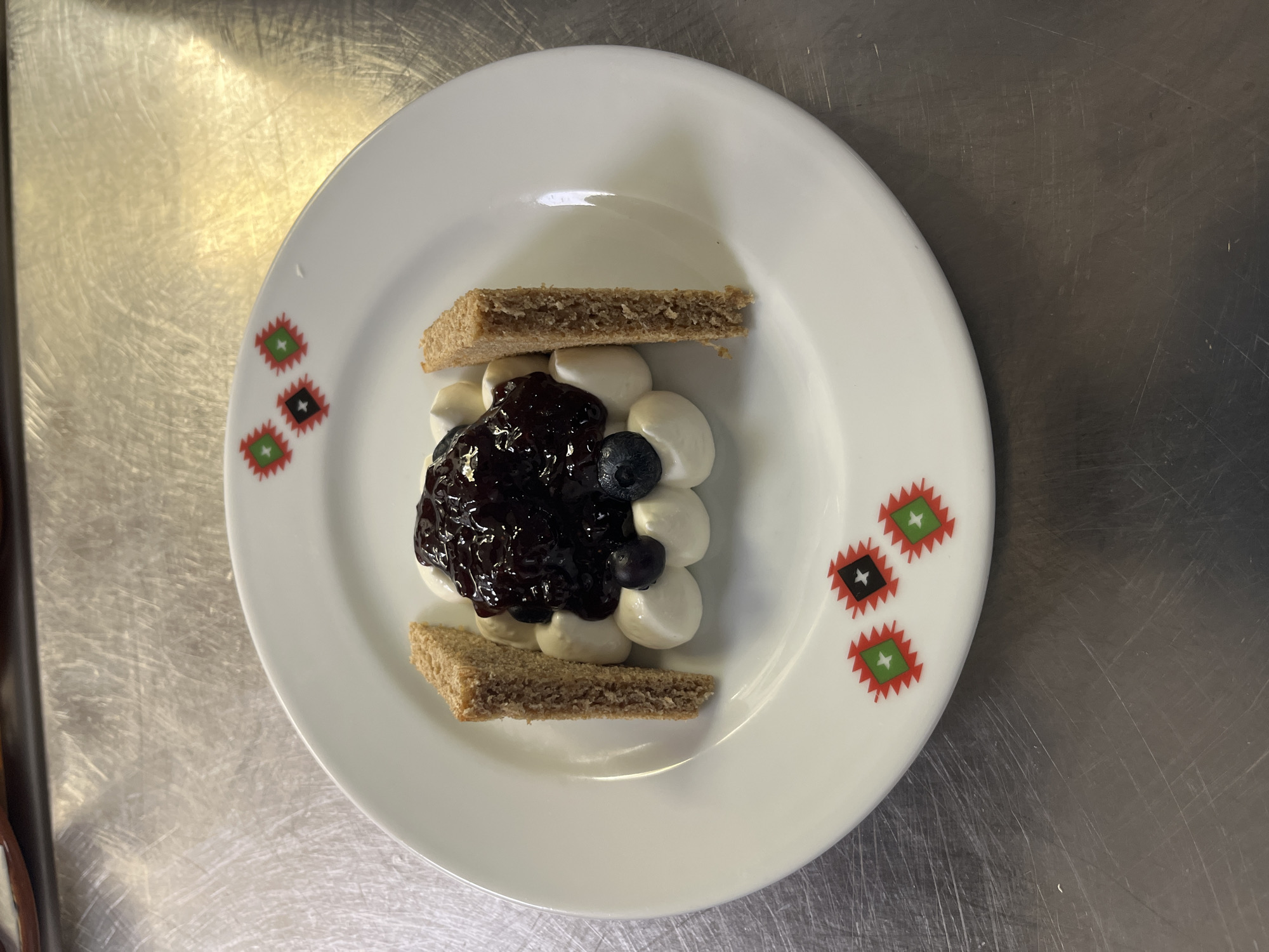 A dessert is presented on a white plate with a decorative rim featuring a repeating geometric pattern of red, green, and black diamonds with white stars. The dessert consists of a dollop of whipped cream topped with dark berry compote, garnished with whole blueberries. Two rectangular pieces of light brown cake, possibly biscotti or a thin slice of sponge cake, are placed vertically on either side of the whipped cream mound. The plate rests on a stainless steel surface, which appears to be a countertop or table, with visible scratches and reflections. The lighting suggests an indoor environment. The image does not contain any information or visual cues that would allow for the determination of a specific location within the city of Varna, Bulgaria.