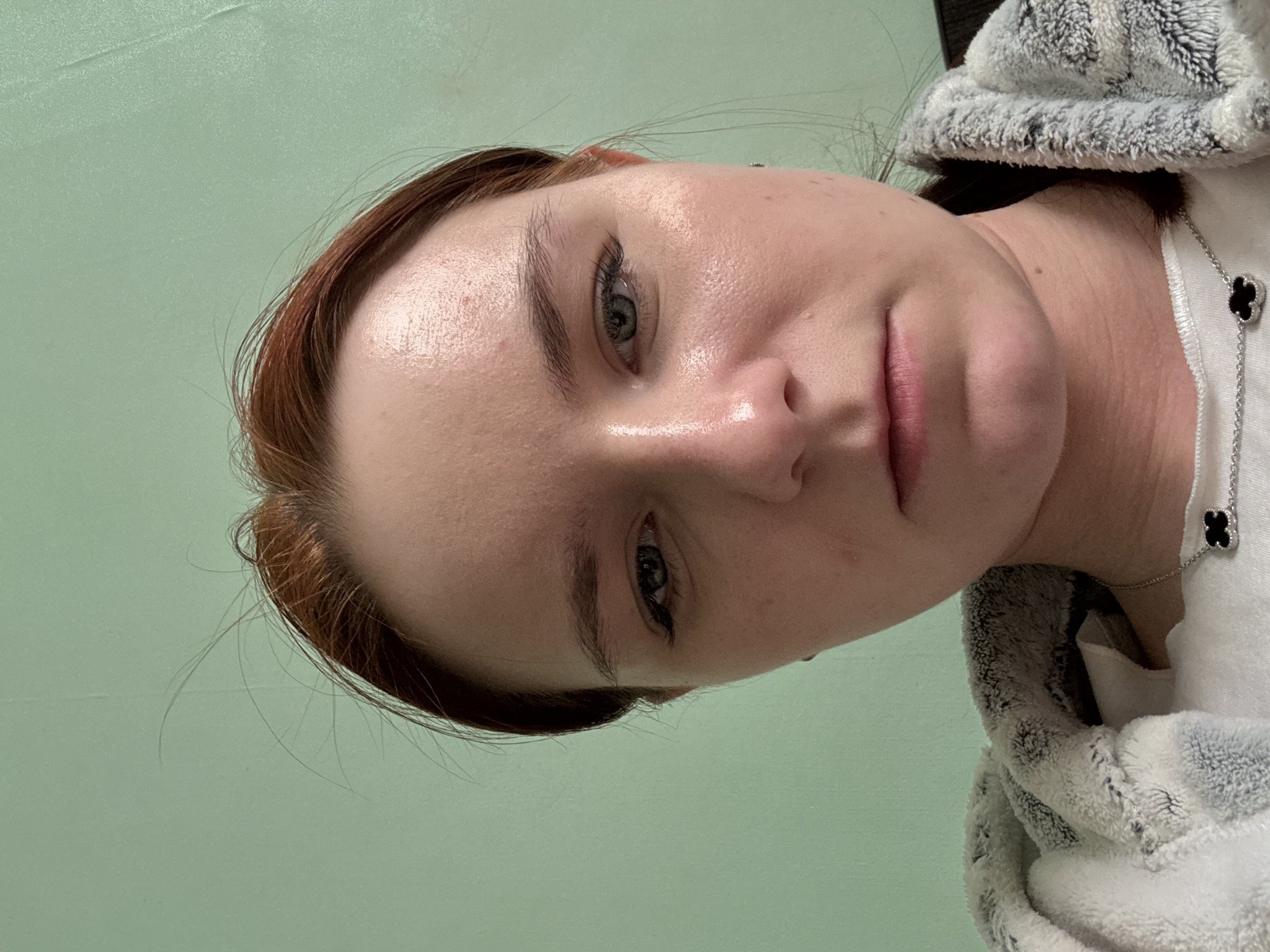 A close-up, slightly angled headshot of a woman with fair skin, light eyes, and reddish-brown hair. She appears to be wearing a white top and a gray and white fuzzy robe or jacket. A silver necklace with a clover-shaped pendant is visible on her neck. The background is a solid light green color. There is no information to place this scene in Saint Petersburg, Russia, or any other specific location.
