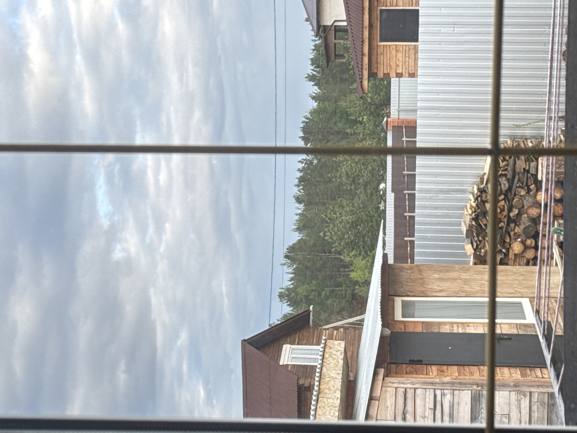 The image shows a view through a window with multiple panes. The outside view features a cloudy sky and a landscape containing houses and trees. In the middle ground, there's a dense forest of green trees. A house is visible on the right side, constructed with wood and metal siding. There is also a pile of logs stacked against the house. Another wooden house is visible on the lower left, with a metal roof. Power lines are seen across the upper portion of the scene, against the sky.
