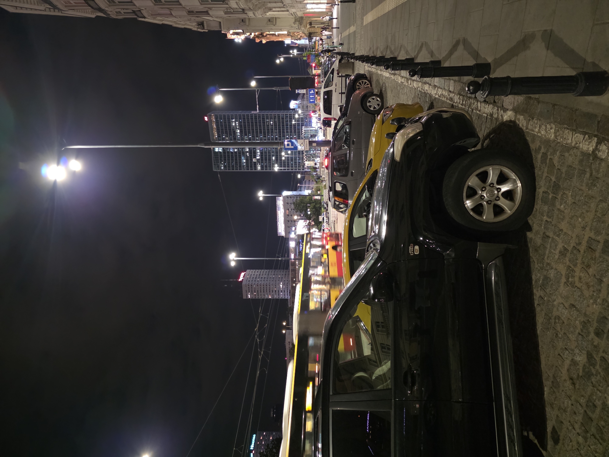 The image depicts a nighttime scene on a street in Warsaw, Poland. The foreground is dominated by the side of a black SUV, parked on a cobblestone street. Several other cars, including a yellow sedan and a gray SUV, are parked alongside it. In the middle ground, a yellow and red tram or bus is in motion, its lights blurred due to the long exposure. Beyond the vehicle, the street is lined with bollards and a sidewalk. In the background, modern skyscrapers with illuminated windows rise into the dark sky, and streetlights cast pools of light onto the pavement. The scene conveys a sense of urban activity at night.