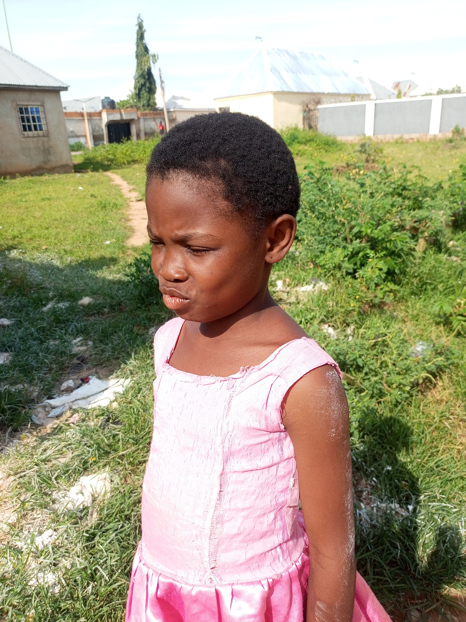 A young girl with dark skin and black hair stands outside, her head turned to the left and her eyes partially closed. She wears a pink dress. The background consists of dry grass, dirt, and some distant buildings with corrugated metal roofs, suggesting a rural or semi-urban setting in Tempure, Nigeria. The lighting indicates it is daytime, likely sunny with shadows cast on the ground.