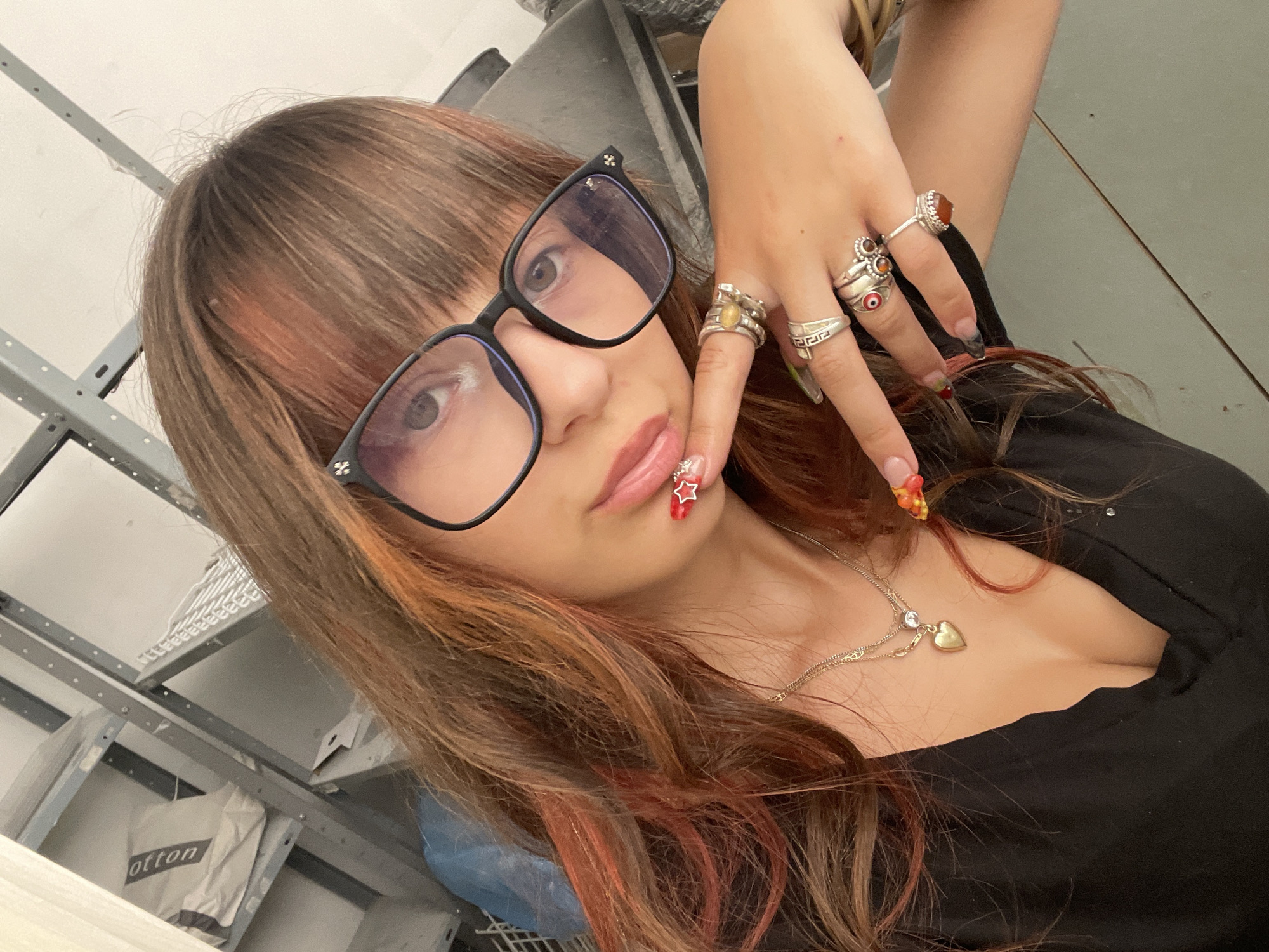 A young woman with long, auburn hair, featuring streaks of red and orange, poses for a selfie indoors. She is wearing large, black-rimmed glasses and a black V-neck top. Her right hand is raised, with fingers adorned with multiple silver rings and intricate nail art. The background appears to be a storage area or workshop, with grey metal shelving units visible. The shelving units contain various items, including bags and what appear to be metal components. The overall lighting suggests an indoor environment, possibly a garage or storage unit. The image is taken from a slightly elevated angle, looking down at the woman. There is no indication that this scene is located in Raanana, Israel.