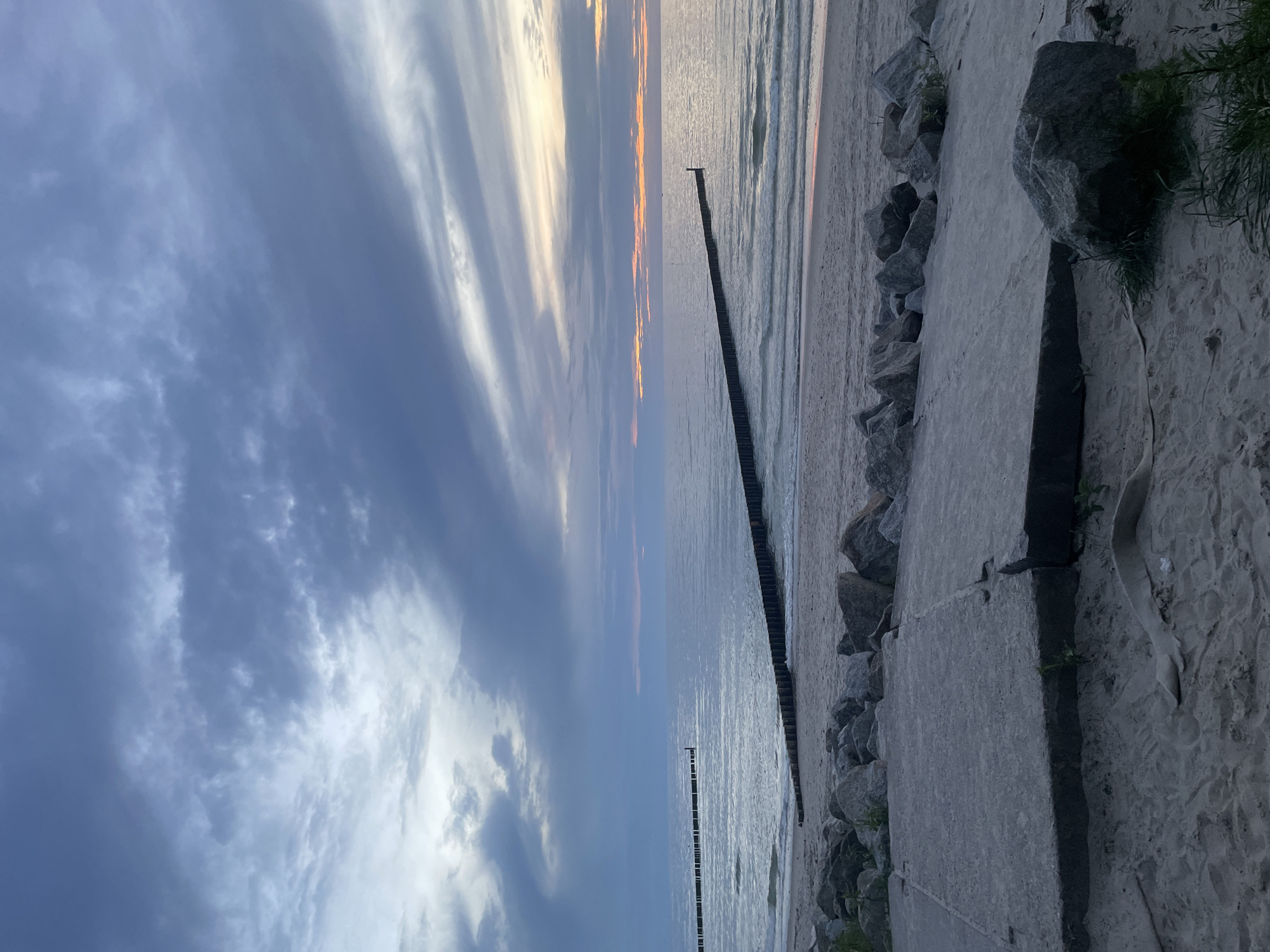 This image depicts a tranquil scene at the beach in Kołobrzeg, Poland, near sunset. The sky is overcast with dramatic clouds in shades of gray and hints of pink and orange from the setting sun, creating a soft, diffused light. The sea is calm, with gentle waves lapping at the sandy shore. Two lines of wooden groins, characteristic of the Baltic coast, extend out into the water, breaking the monotony of the sea. The foreground is occupied by a sandy beach with scattered rocks and a concrete structure, possibly part of a walkway or old pier. The overall atmosphere is serene and peaceful, with no people or animals visible.