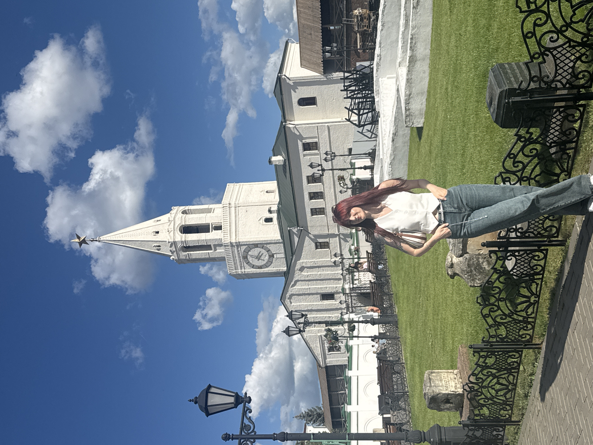 A woman with long, red hair and a white vest stands in front of the Syuyumbike Tower in Kazan Kremlin, Russia. She is wearing blue jeans and a silver chain belt. She has her hands in her pockets and is looking to her left. The Syuyumbike Tower is a tall, white building with a clock face and a star on top. Behind the tower are other buildings with white walls and green roofs. The sky is blue with white clouds. The ground is paved with brick in the foreground and covered with green grass in the background. There is a black metal fence in front of the grass. Several benches and lampposts are visible in the scene.