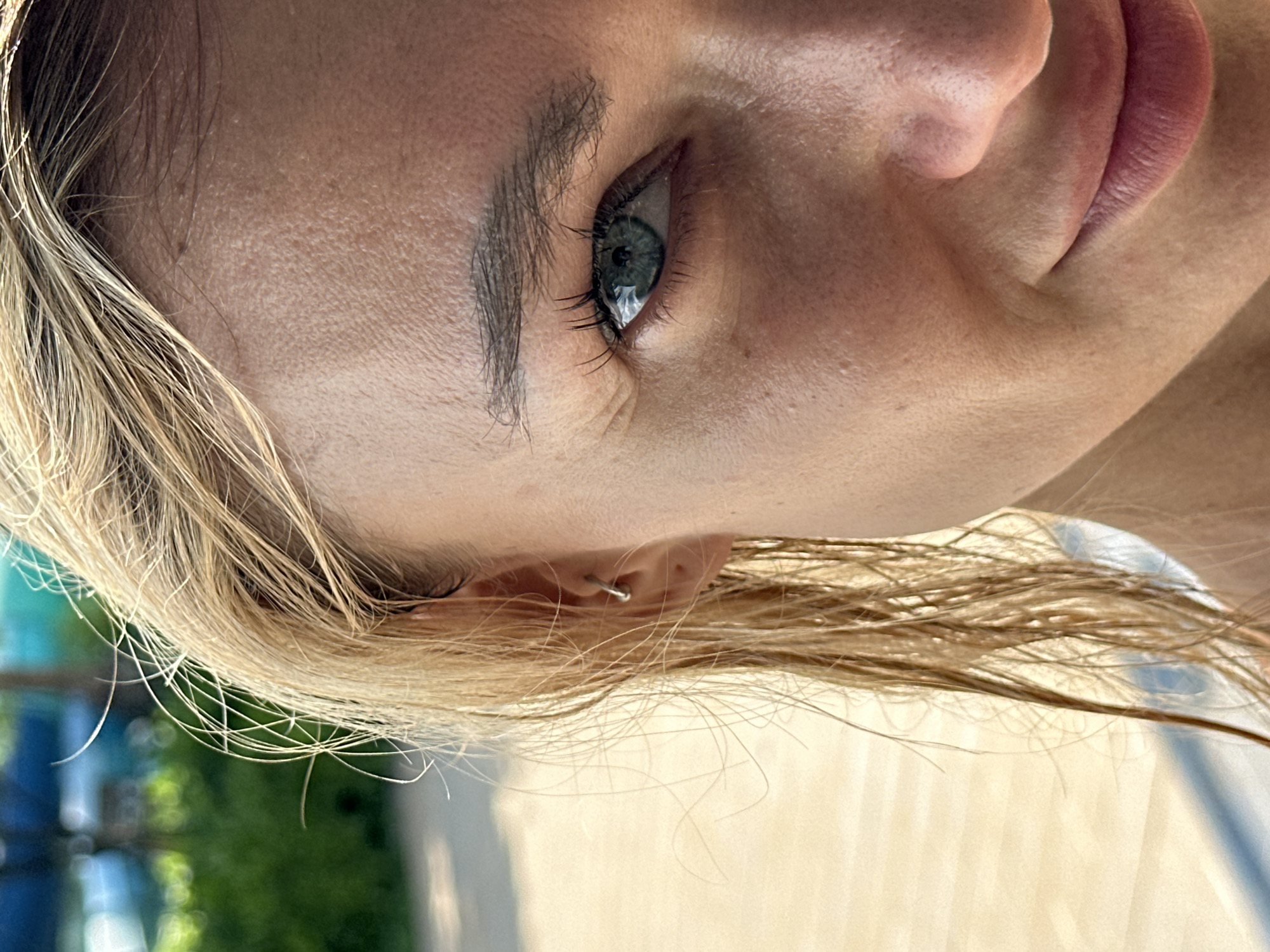 A close-up, right-sided portrait features a fair-skinned female subject with blonde hair, captured outdoors under bright natural light in Tien Thanh Ward, Vietnam. The image focuses on the right side of her face, prominently displaying her right eye, eyebrow, ear, and the right portions of her nose and mouth. Her blonde hair, appearing wet or damp, frames her face, with individual strands and flyaways clearly discernible. A small silver-colored hoop earring is visible in the helix piercing of her right ear. Her right eye exhibits a blue-green iris with a visible pupil and dark, defined eyelashes, directed straight at the viewer. Fine lines are present around the outer corner of her eye. The subject's skin displays visible pores, subtle textures, and highlights from the strong illumination. The right corner of her mouth shows a slight upward curve, indicative of a faint smile. The background is significantly blurred, featuring soft green and blue hues consistent with foliage and water features, alongside lighter, horizontal patterns suggesting a wooden deck or similar outdoor surface, all brightly lit by sunlight.