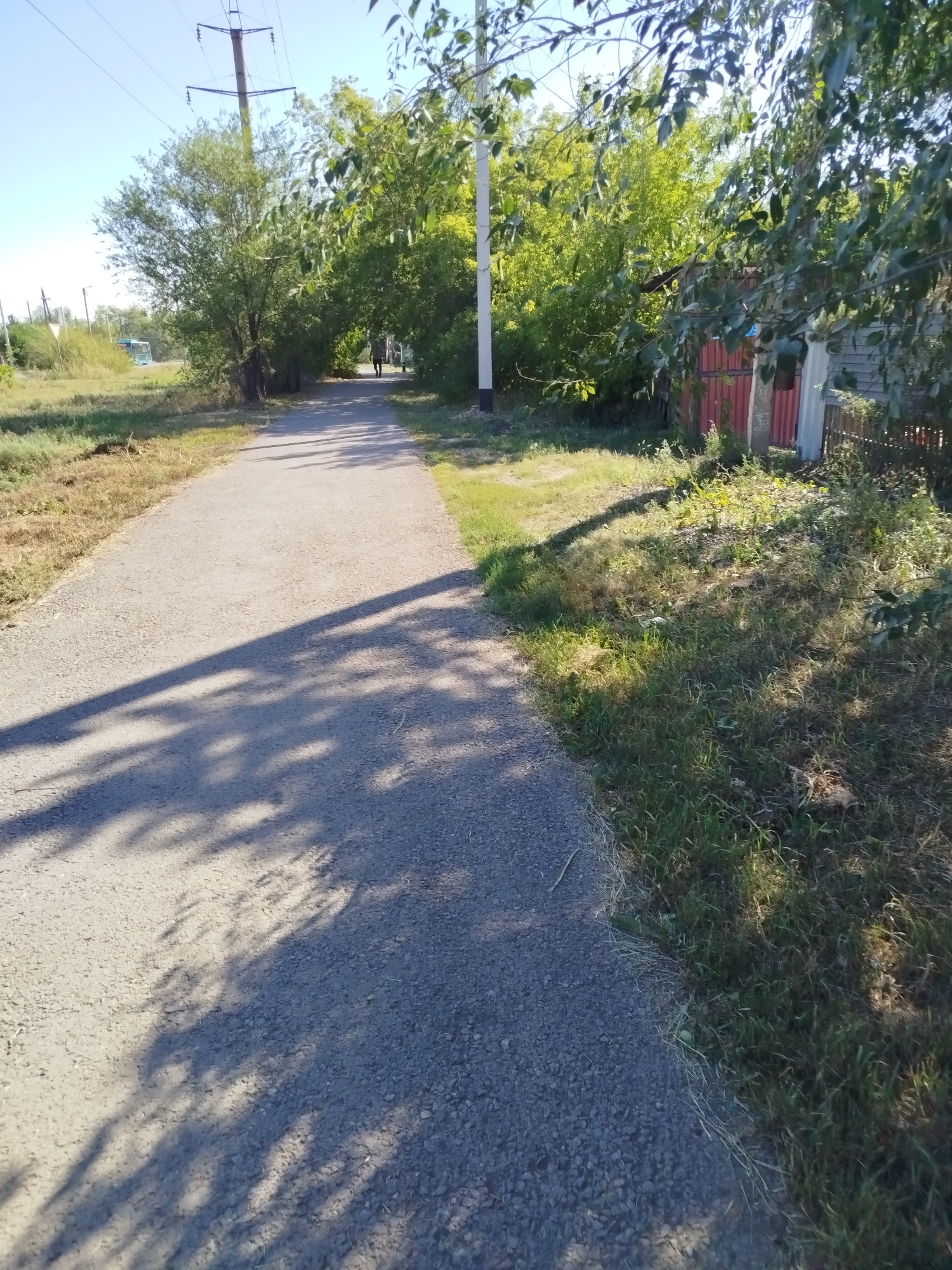 The image depicts a paved pathway lined with vegetation on either side, suggesting a park or a rural setting. The pathway curves gently into the distance, where a lone figure is walking. Trees with green foliage flank the path, casting dappled shadows on the ground. On the right side of the path, a red metal fence and a wooden structure are visible, partially obscured by foliage. Overhead, power lines and poles are present. The sky is clear and blue. The scene appears to be from a daytime setting, likely outdoors, with the sun casting shadows from the trees.