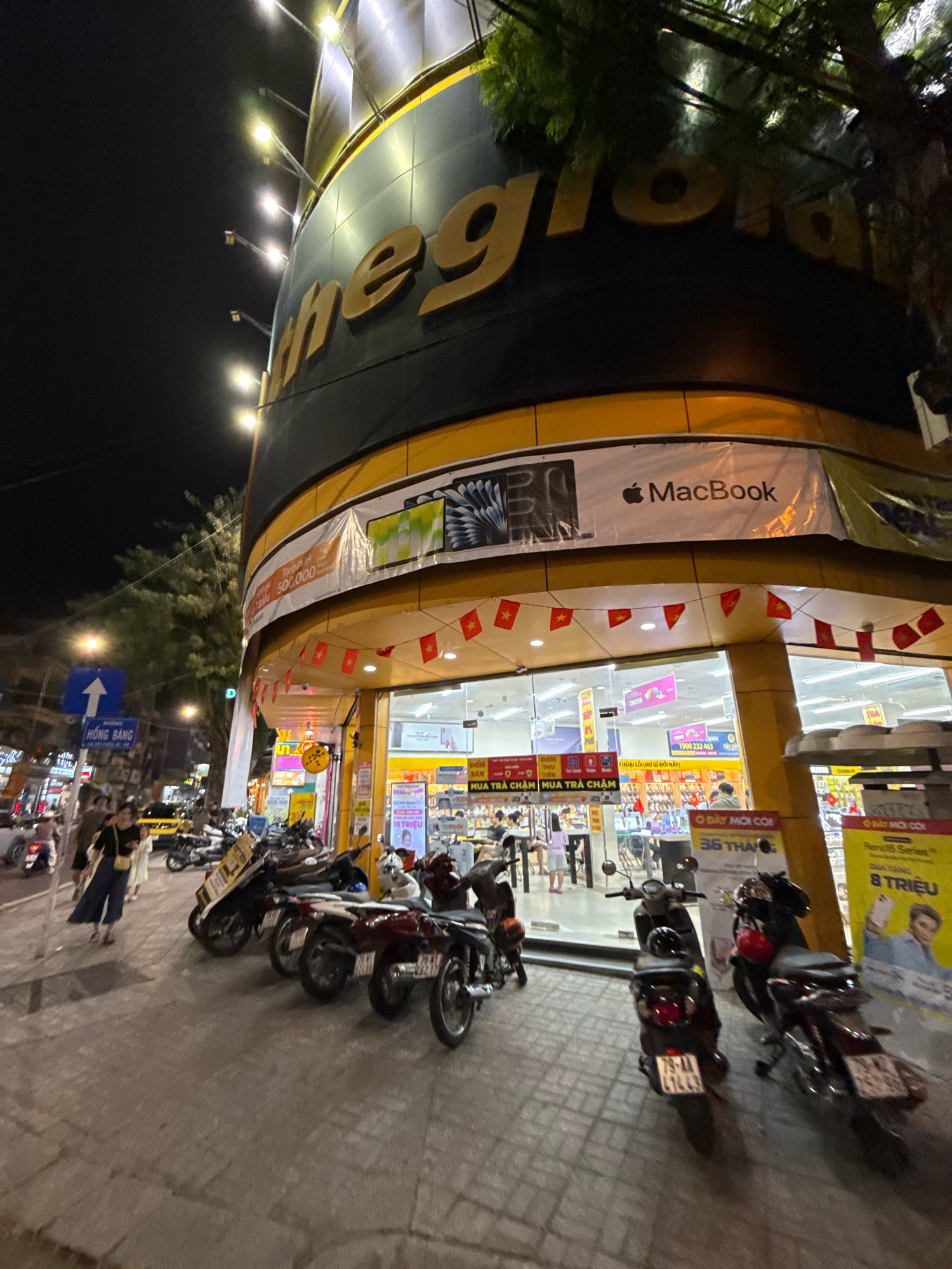 The image shows a nighttime view of a "thegioididong" electronics store in Nha Trang, Vietnam. The store features a large, curved facade with prominent yellow and black signage, including the "thegioididong" logo in gold letters. Above the entrance, a banner advertises "MacBook" with the Apple logo. Red and yellow Vietnamese flags are strung along the exterior.
In front of the store, several motorcycles are parked on the sidewalk and street. A few people are visible, including a woman in a dark shirt and blue pants with a yellow bag walking on the sidewalk, and other individuals inside and outside the store. The street is illuminated by streetlights and the store's interior lighting. A blue traffic sign with a white arrow and Vietnamese text is visible on the left side of the image. The overall scene depicts a typical urban streetscape at night in Vietnam, focusing on a retail environment.