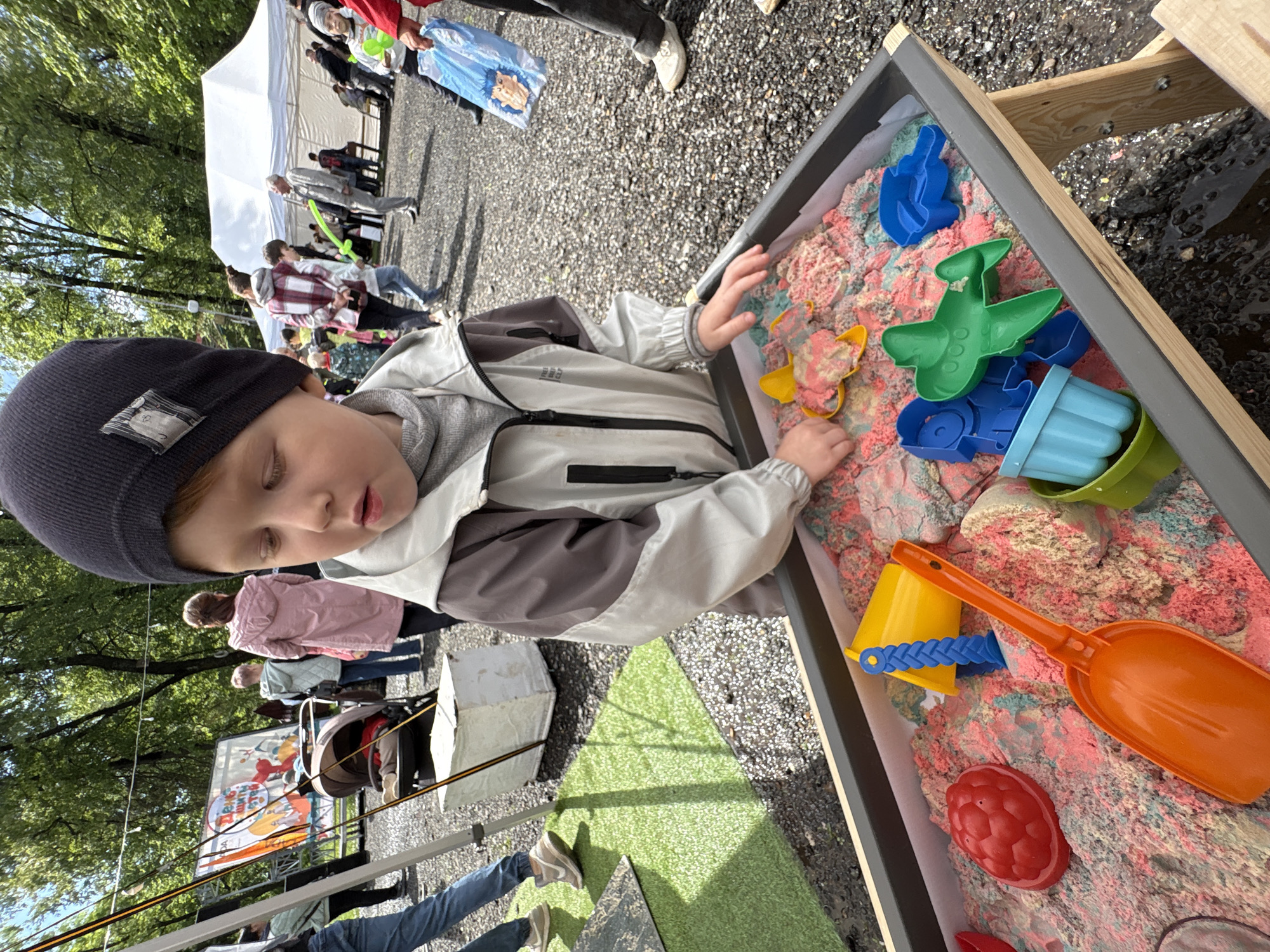 A child wearing a navy blue knit hat and a gray and brown jacket is playing with toys in a sand table. The sand table is filled with colorful sand and various plastic toys, including a yellow butterfly mold, a green airplane mold, a blue hand mold, a blue and light green pail, a small yellow bucket with a blue handle, an orange shovel, and a red berry-shaped mold. The sand table is located outdoors on gravel. In the background, there are people walking around, a white tent, trees, a promotional banner, and a stroller. No evidence to support the location as Izhevsk, Russia.