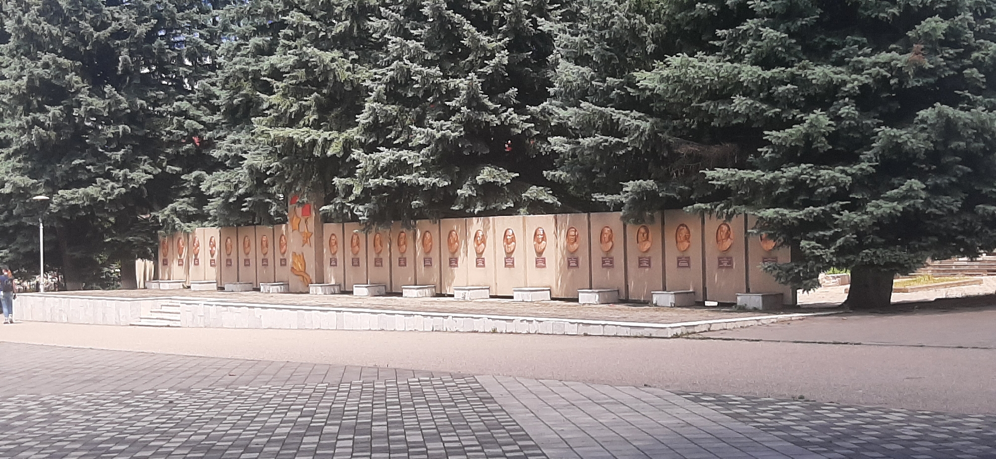 In Yessentukskaya, Russia, a memorial wall lined with busts of notable individuals stands behind a low concrete barrier with steps leading up. The wall is adorned with numerous circular bronze busts, each set against a pale, textured background. Below each bust is a rectangular plaque. The memorial is backed by a dense row of tall, mature evergreen trees, their branches creating a dark canopy. In the foreground, a patterned paved area extends towards the viewer, with a grey asphalt path separating it from the memorial. A single person is visible walking on the left side of the paved area, and a lamppost stands near the trees. The overall scene depicts a public memorial space, likely a park or square, under a bright sky.