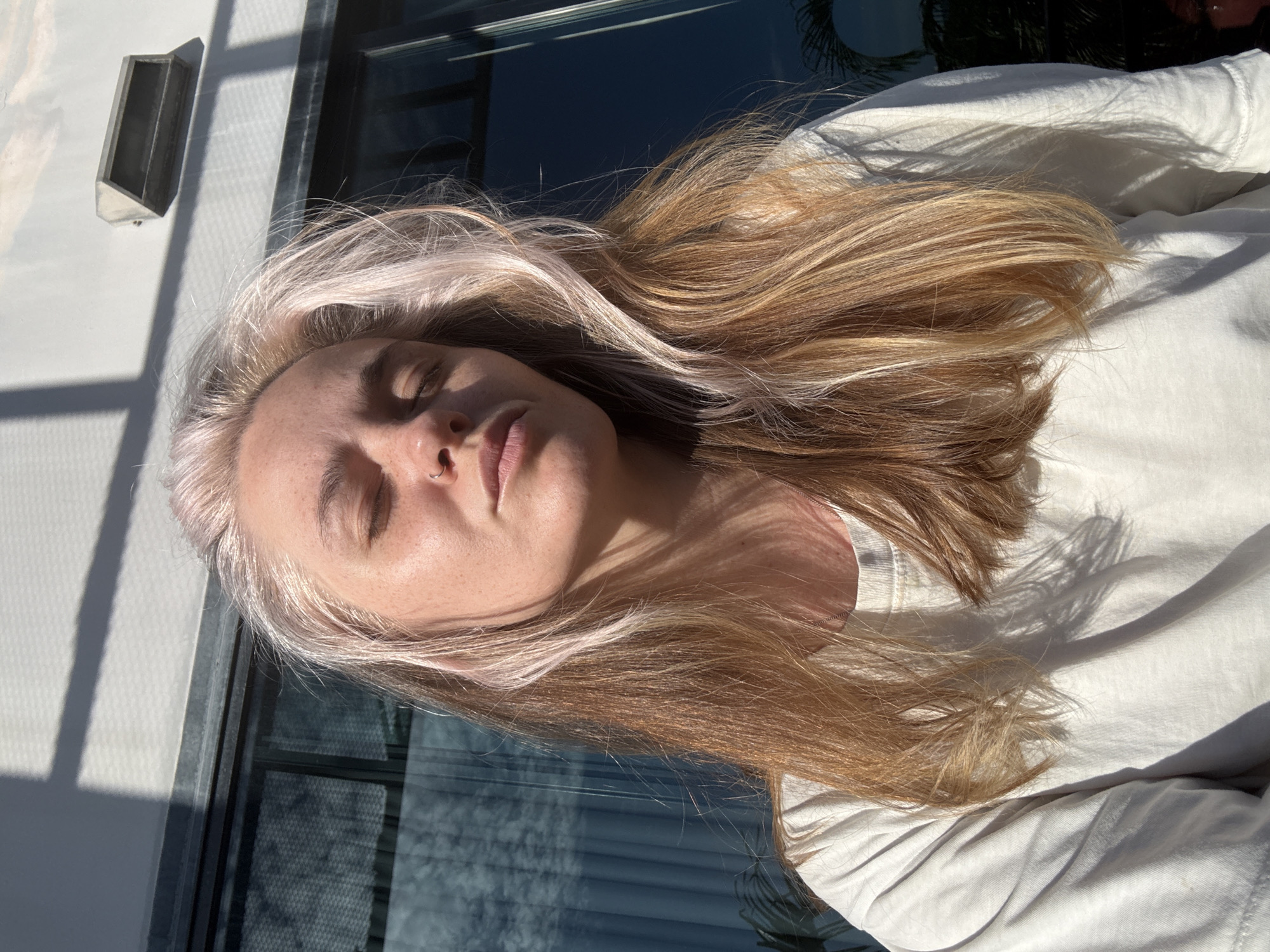 A young woman with blonde and pink-tinged hair is pictured outdoors. She is wearing a white t-shirt and has a nose ring. Her eyes are closed, and her face is tilted upwards, catching the sunlight. The background features a building with large windows and a vent on the wall. Shadows from the sun are cast across the scene, indicating it is daytime. The environment suggests an urban or suburban setting, possibly a residential or commercial area.