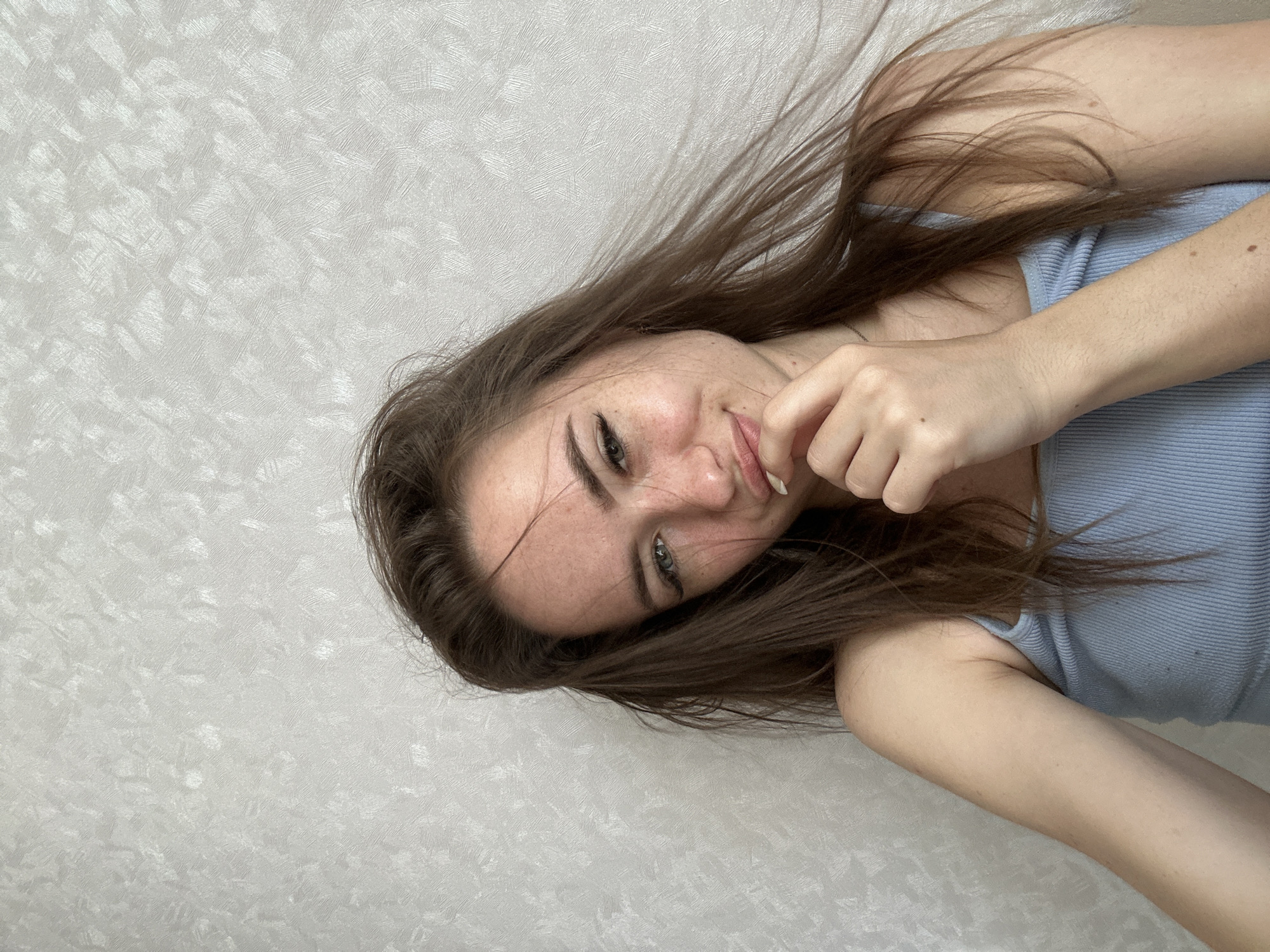 A young person, likely female, with long, dark brown hair is depicted in a close-up, rotated image. She is lying down, looking towards the camera with a slight smile or smirk. Her body is oriented horizontally across the frame, suggesting a relaxed, reclined pose against a light, textured wall.
The subject is wearing a light blue ribbed tank top or camisole. Her hair is spread out, flowing around her head and shoulders, with some strands falling across her face. One hand is raised, with fingers gently touching her chin or lower lip, partially obscuring her mouth.
Notable details include a scattering of freckles visible across her nose and cheeks. Her eyes are light in color, possibly blue or green, and she appears to be wearing eyeliner. Her complexion is fair, and several small moles are visible on her arms. The background is a uniform, light gray or off-white wall with a subtle, abstract textured pattern, which appears to be wallpaper or painted plaster. The lighting is soft and even, suggesting an indoor setting. There is no visible text in the image.