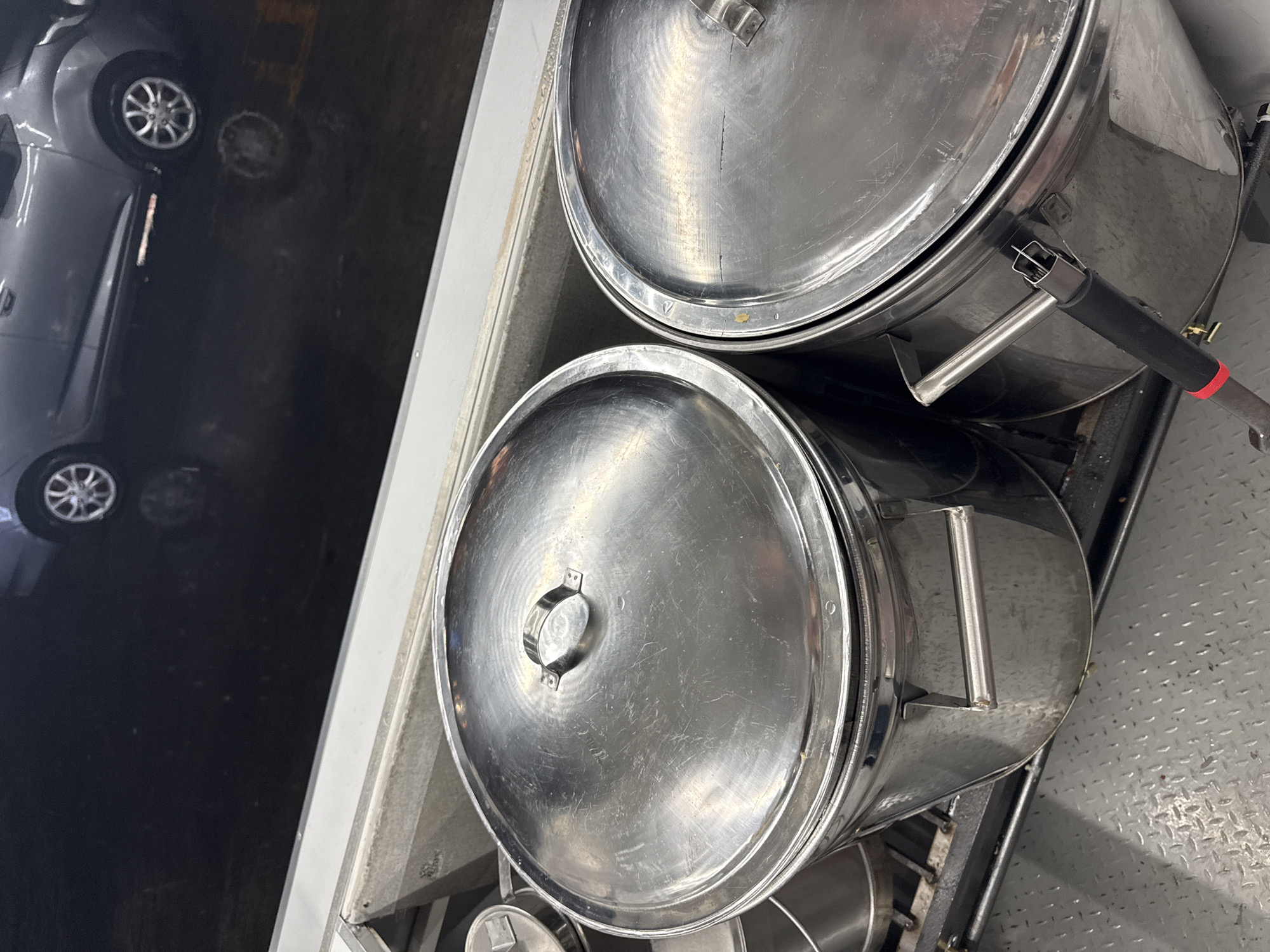 The image shows two large, stainless steel cooking pots with lids, stacked on a metal rack. One pot has a long handle attached. To the left of the pots is what appears to be the front of a silver car parked on a dark surface, potentially asphalt. The scene is oriented vertically. The location is not evident from the image and cannot be determined.
